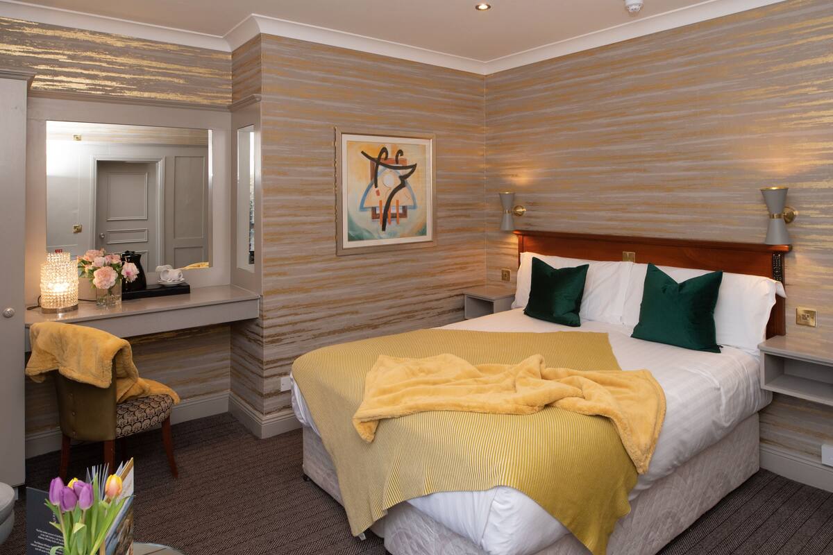 A hotel room with a bed, a desk, a chair, a mirror, and a painting on the wall.
