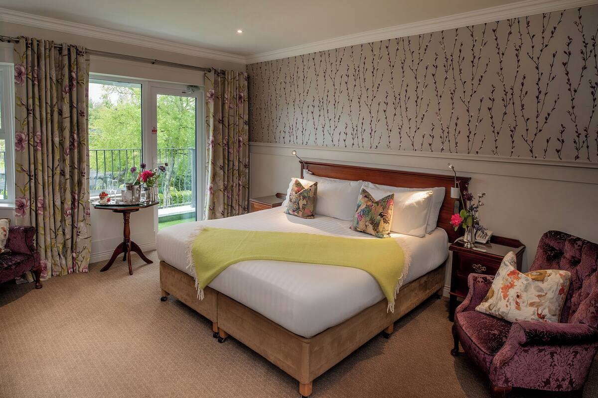 A bedroom with a large bed, floral curtains, a small table with flowers, and a view of greenery through the window.