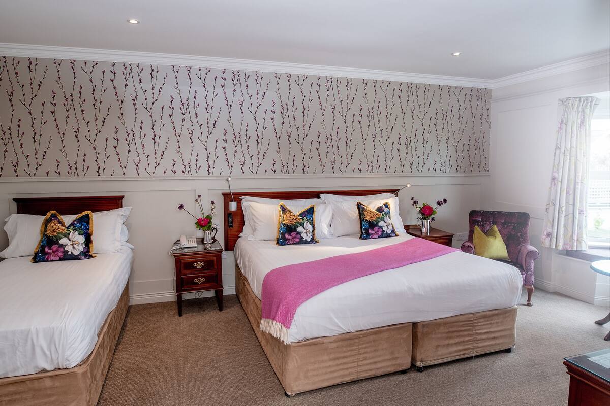 A hotel room with two beds, a large window with curtains, and a floral wallpaper design.