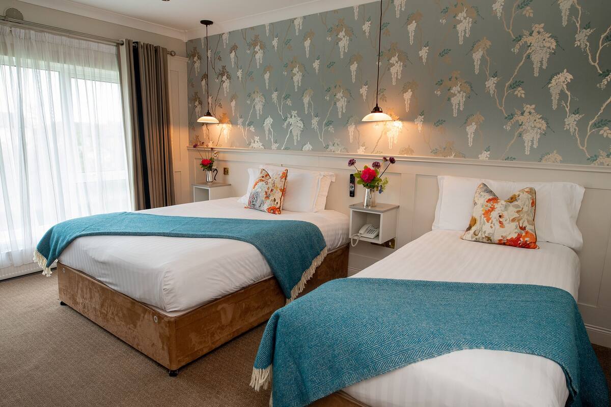 A hotel room with two beds, a large window with curtains, floral wallpaper, and bedside lamps.