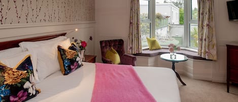 A hotel room with a large bed, floral-patterned pillows, a pink blanket, a round table with a vase, and a view of a garden through a window.