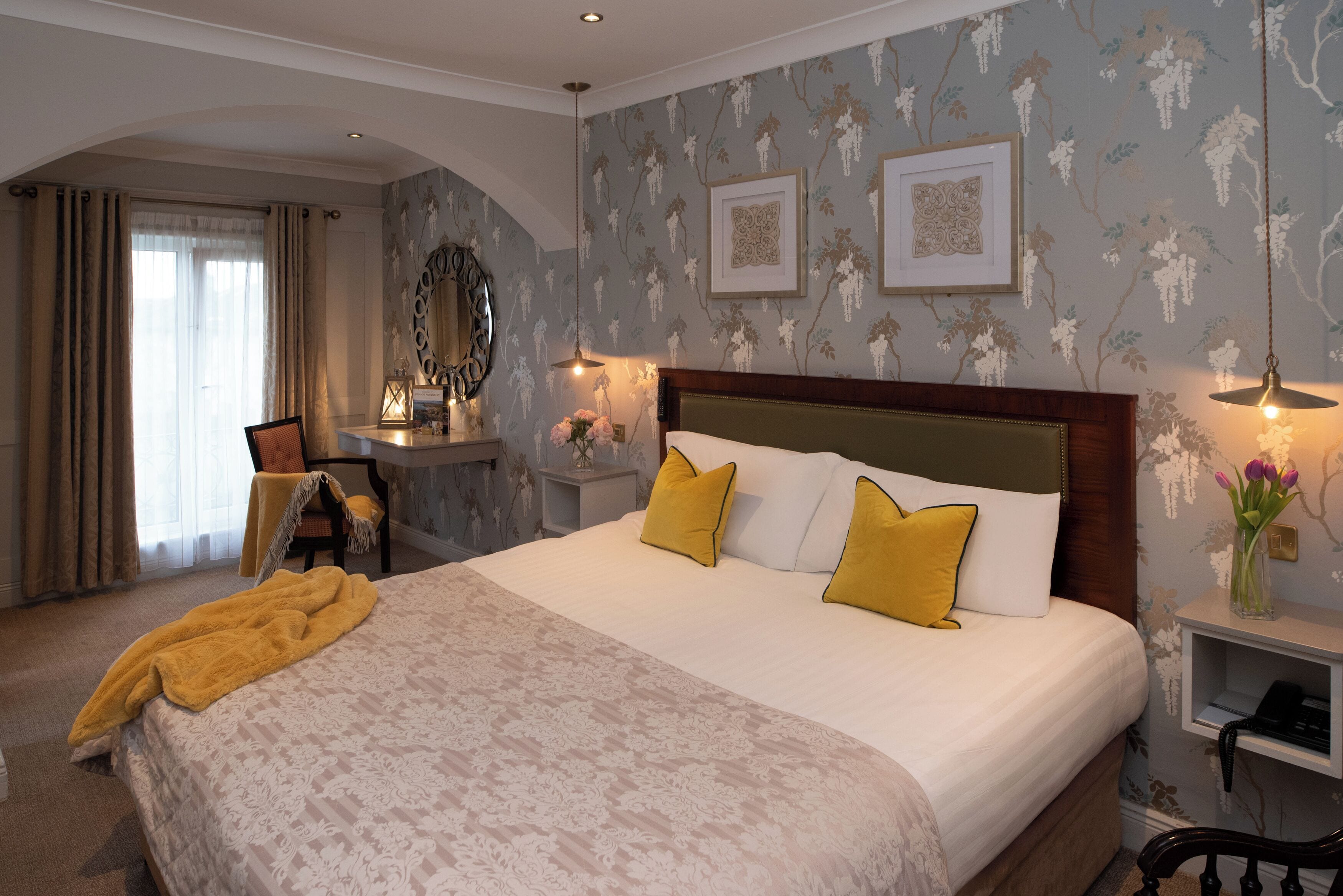 Quarto de hotel com uma cama grande, uma escrivaninha, uma cadeira, um espelho e uma mesa de cabeceira com luminária.