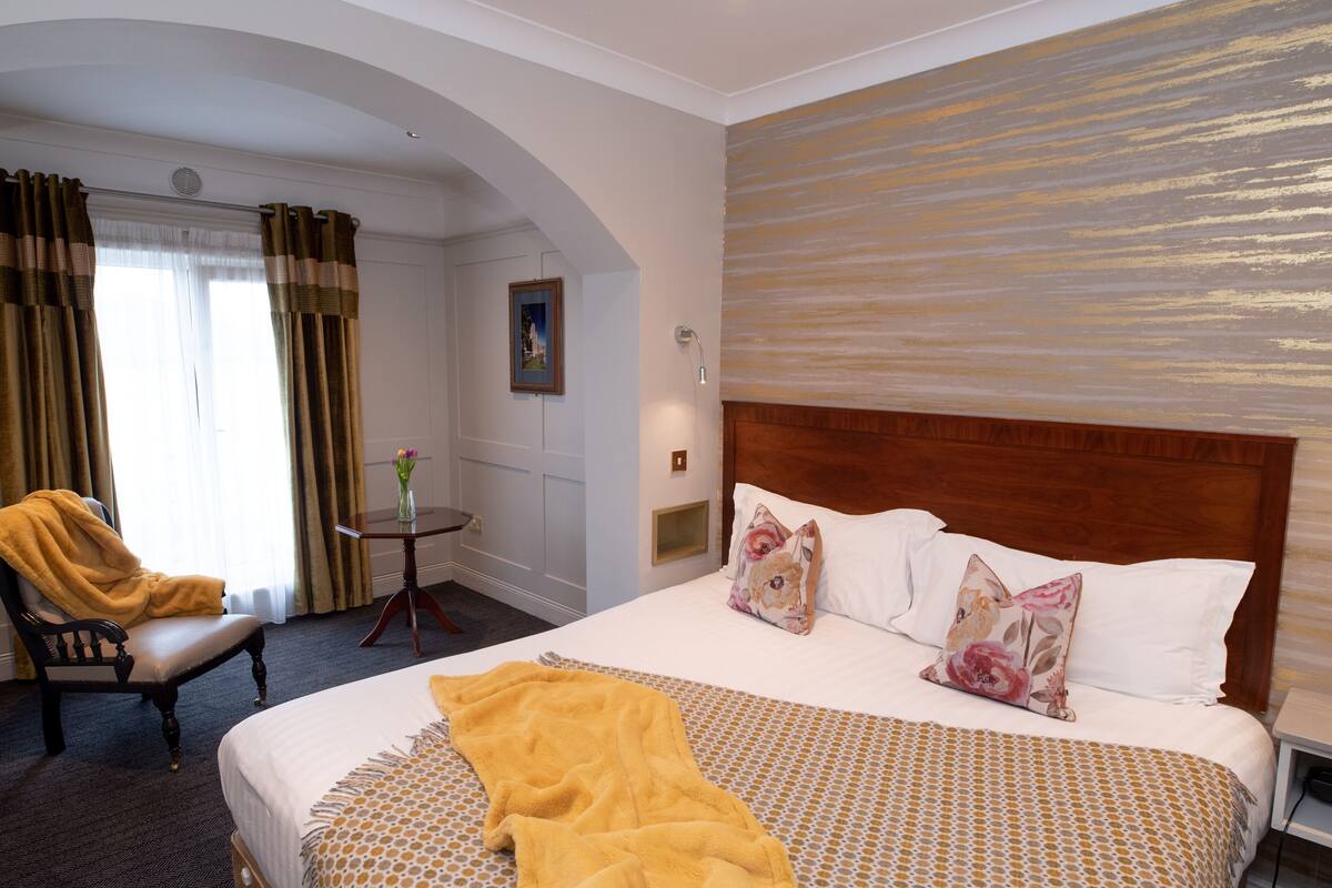 A hotel room with a large bed, a wooden headboard, a small table with a vase, and a chair with a yellow blanket.