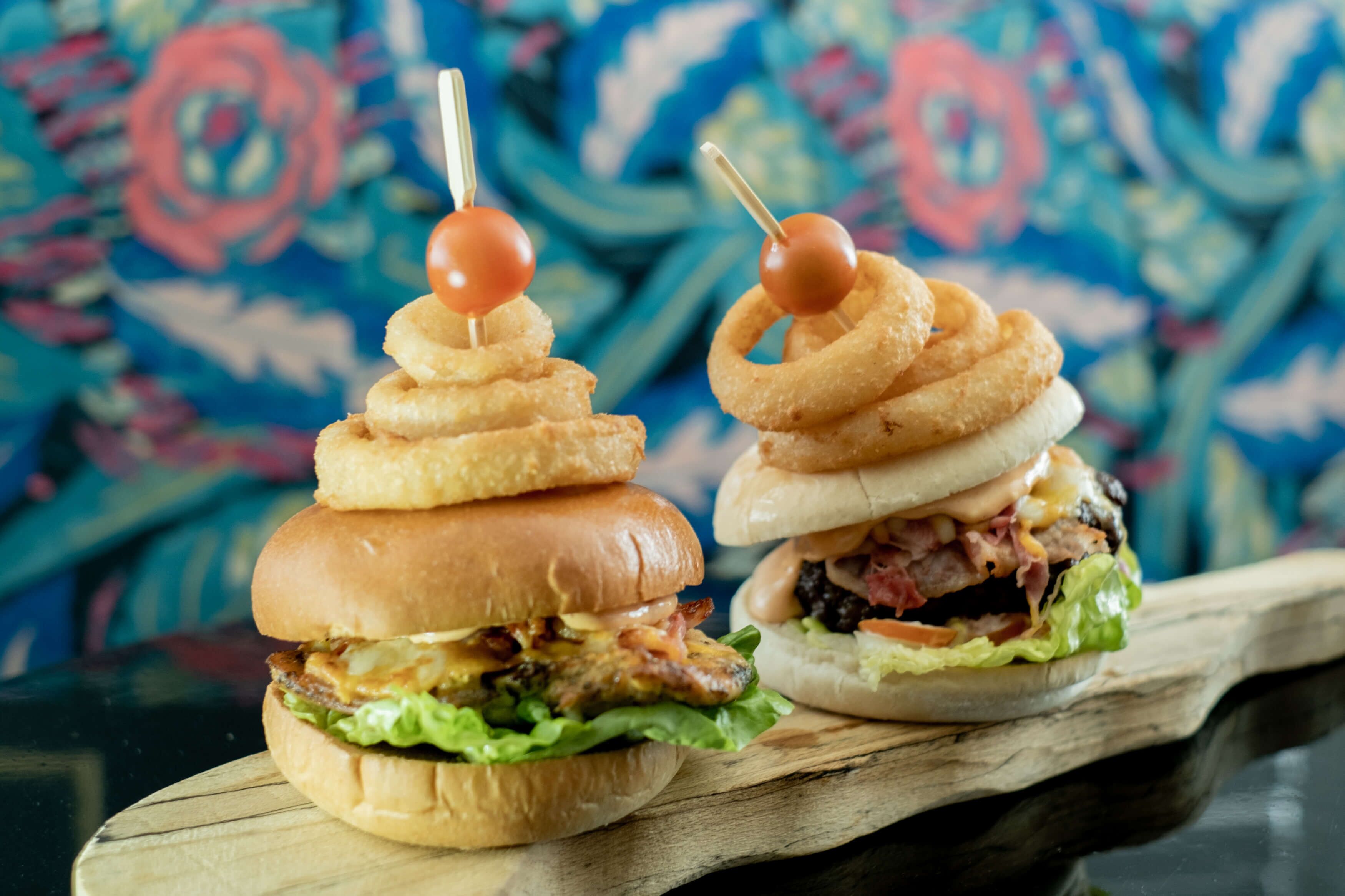 Two burgers with onion rings on skewers, served on a wooden board.