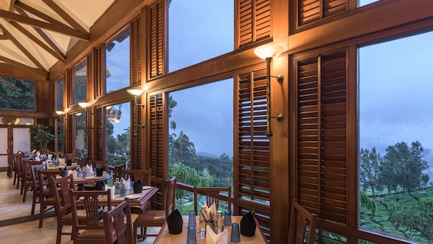 A restaurant with wooden furniture and large windows overlooking a lush landscape. - Club Mahindra Munnar (Devikolam)