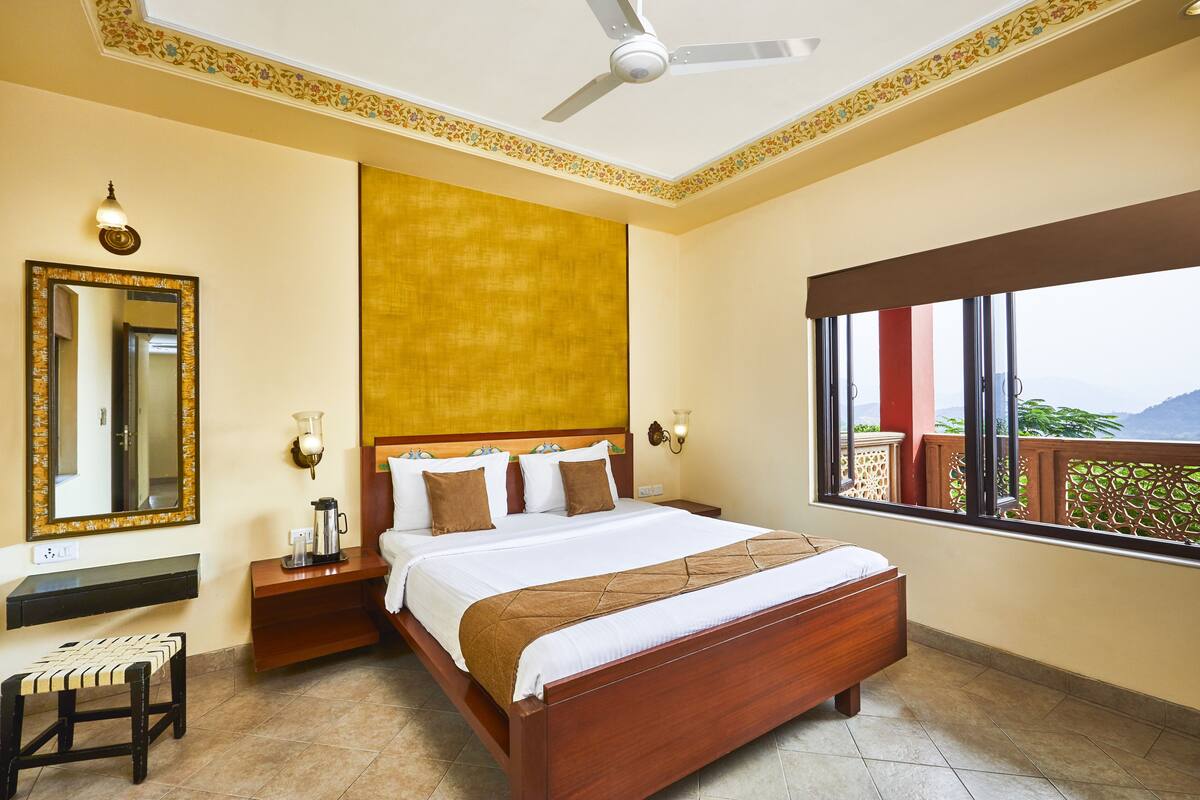 Habitación de hotel con cama, mesita de noche, banco, espejo, ventana con vistas y ventilador de techo.