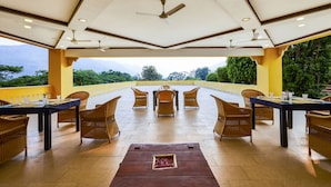An outdoor dining area with wicker furniture, ceiling fans, and a view of greenery and mountains. - Club Mahindra Kumbhalgarh (Rajsamand)