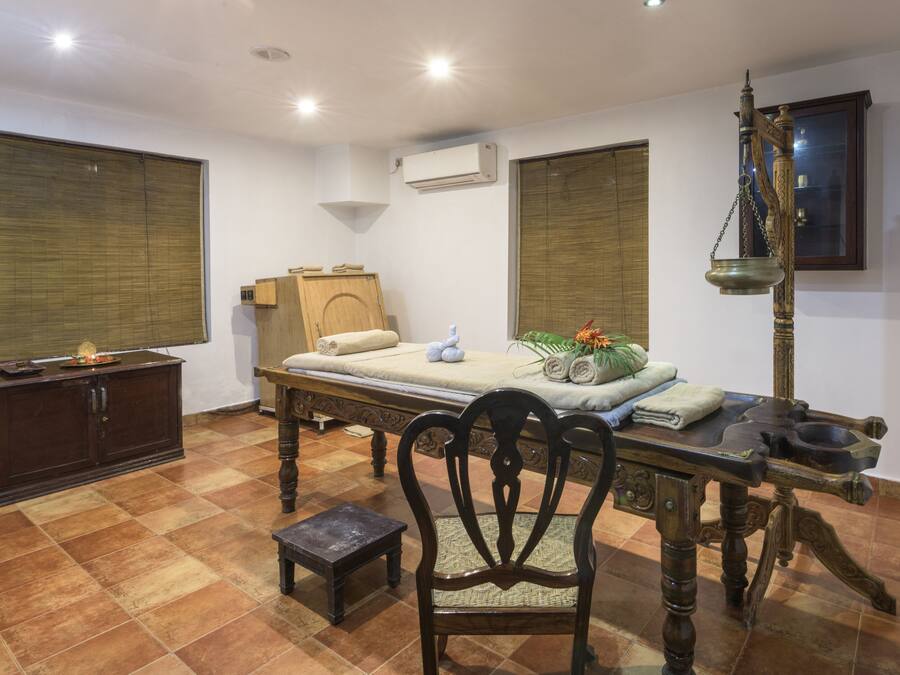A room with a bed, a wooden cabinet, a chair, a table with a lamp, and a window with blinds.