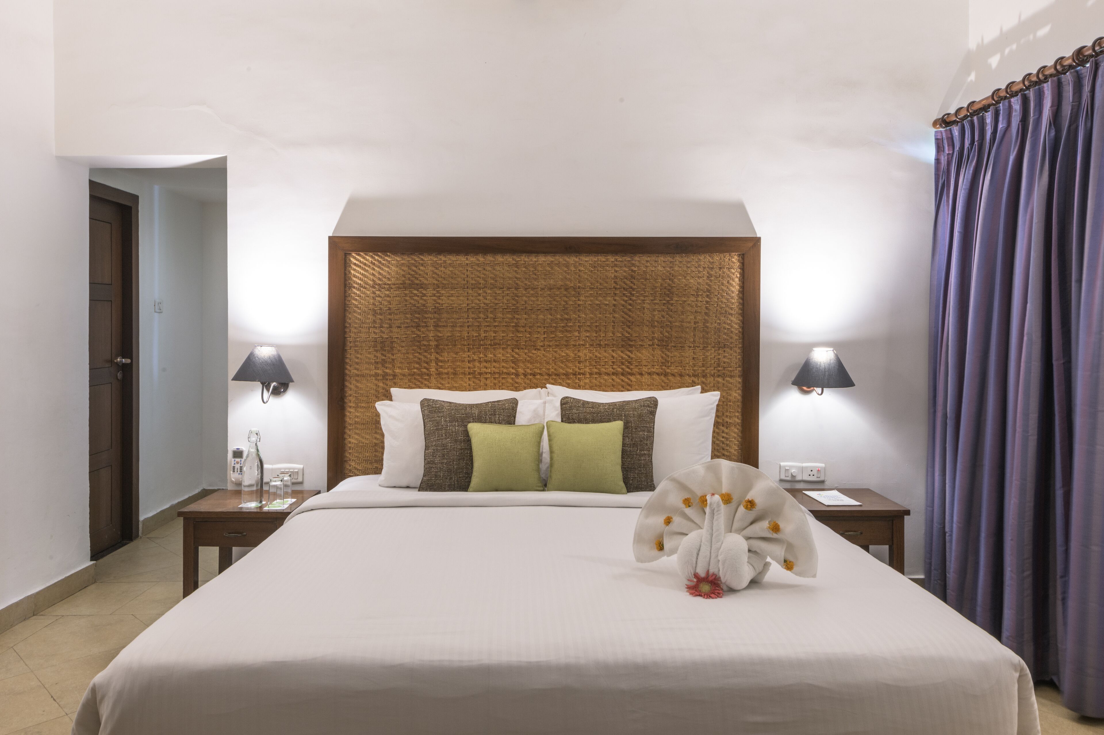 Una cama bien hecha con cabecera, dos lámparas de noche, una mesita con un teléfono y un vaso, y una ventana con cortinas.