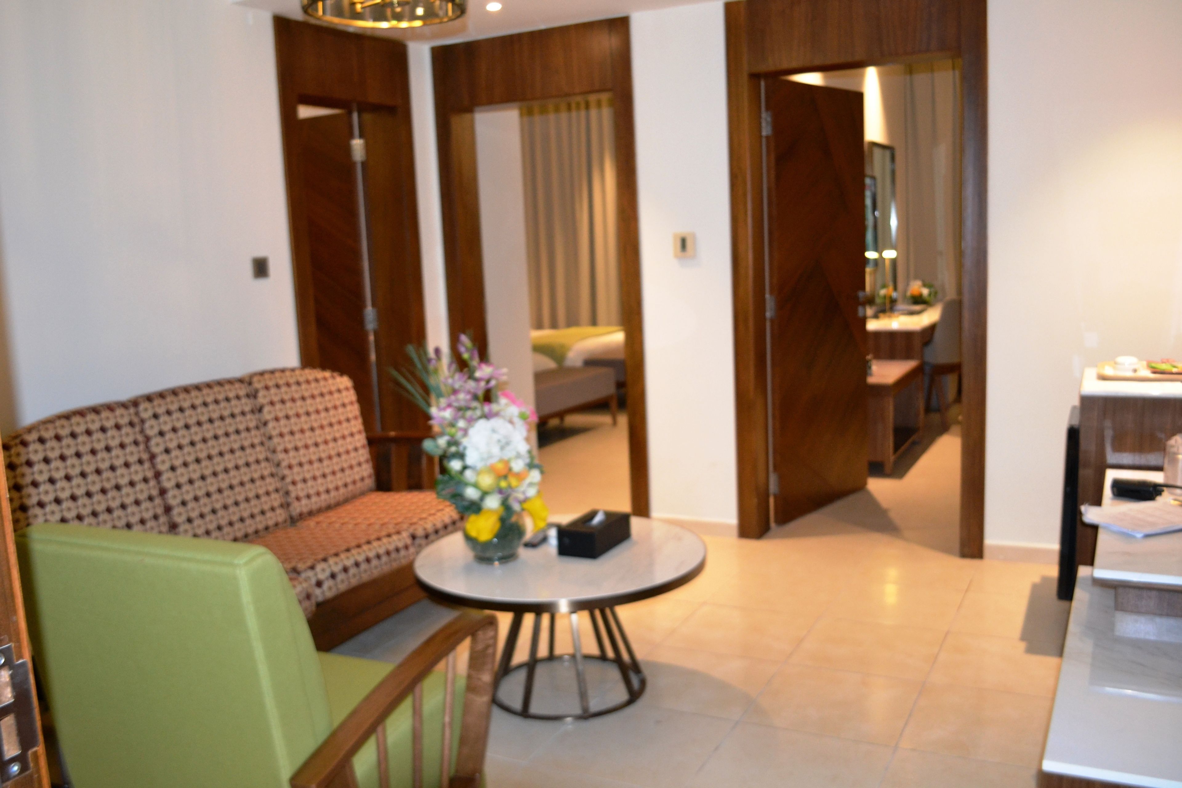 A hotel room with a patterned sofa, a small round table with a vase of flowers, and a wooden door leading to a bathroom.