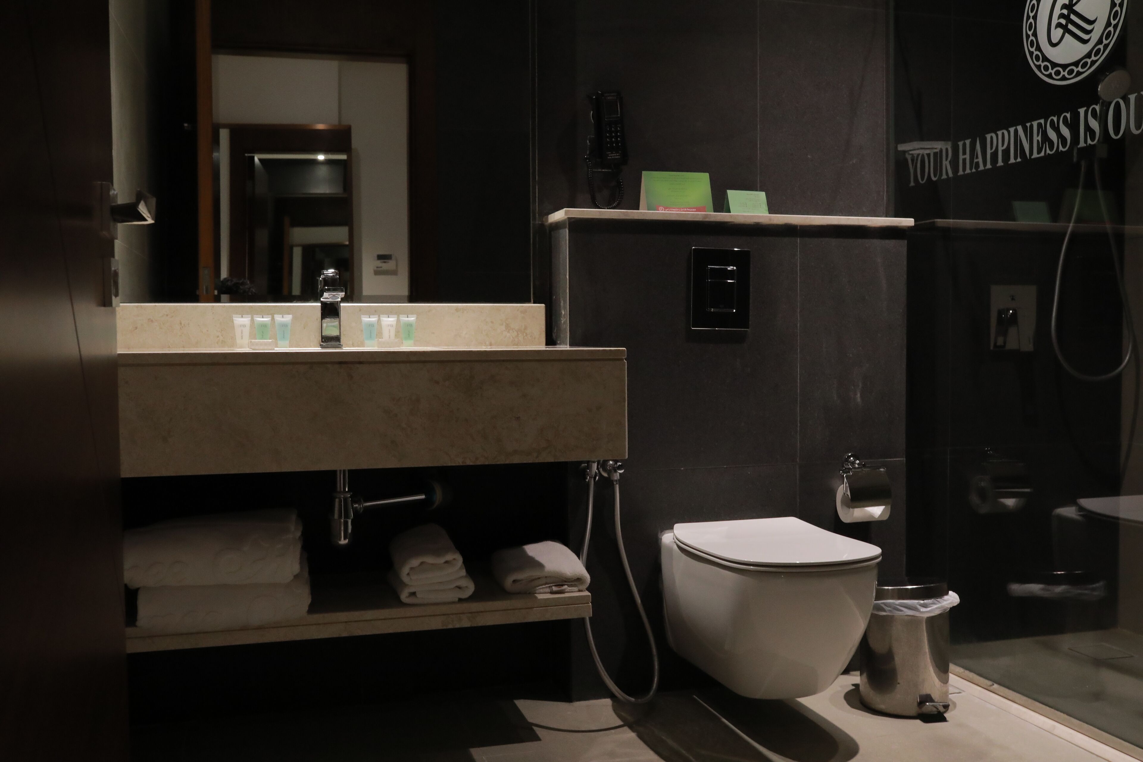 A modern bathroom with a sink, mirror, and toilet. There is a towel rack with folded towels, a shower area, and a sign on the wall.