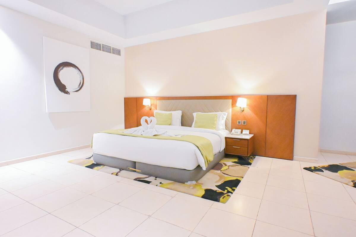 Una habitación de hotel con una cama grande, dos mesitas de noche, un cabecero con lámparas fijadas a la pared y un adorno en la pared.