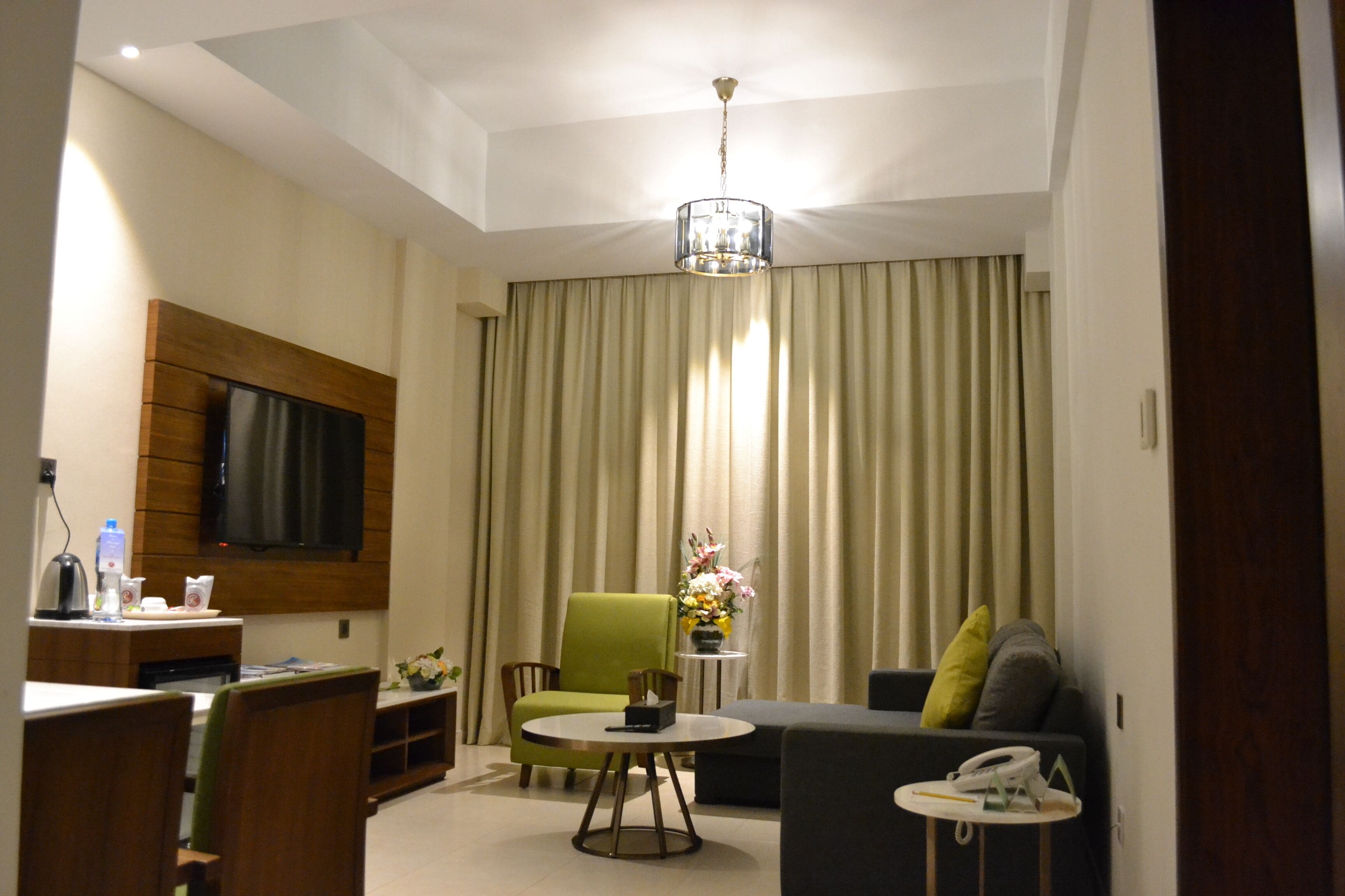 A modern hotel room with a flat-screen TV mounted on the wall, a green armchair, a sofa with cushions, a small round table with a vase of flowers, and a ceiling light fixture.