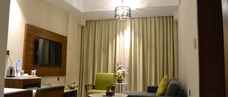 A modern hotel room with a flat-screen TV mounted on the wall, a green armchair, a sofa with cushions, a small round table with a vase of flowers, and a ceiling light fixture.
