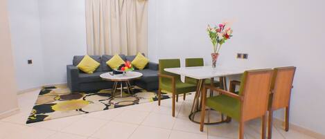 Una sala de estar con un sofá, sillas verdes, una mesa de comedor y un jarrón con flores.