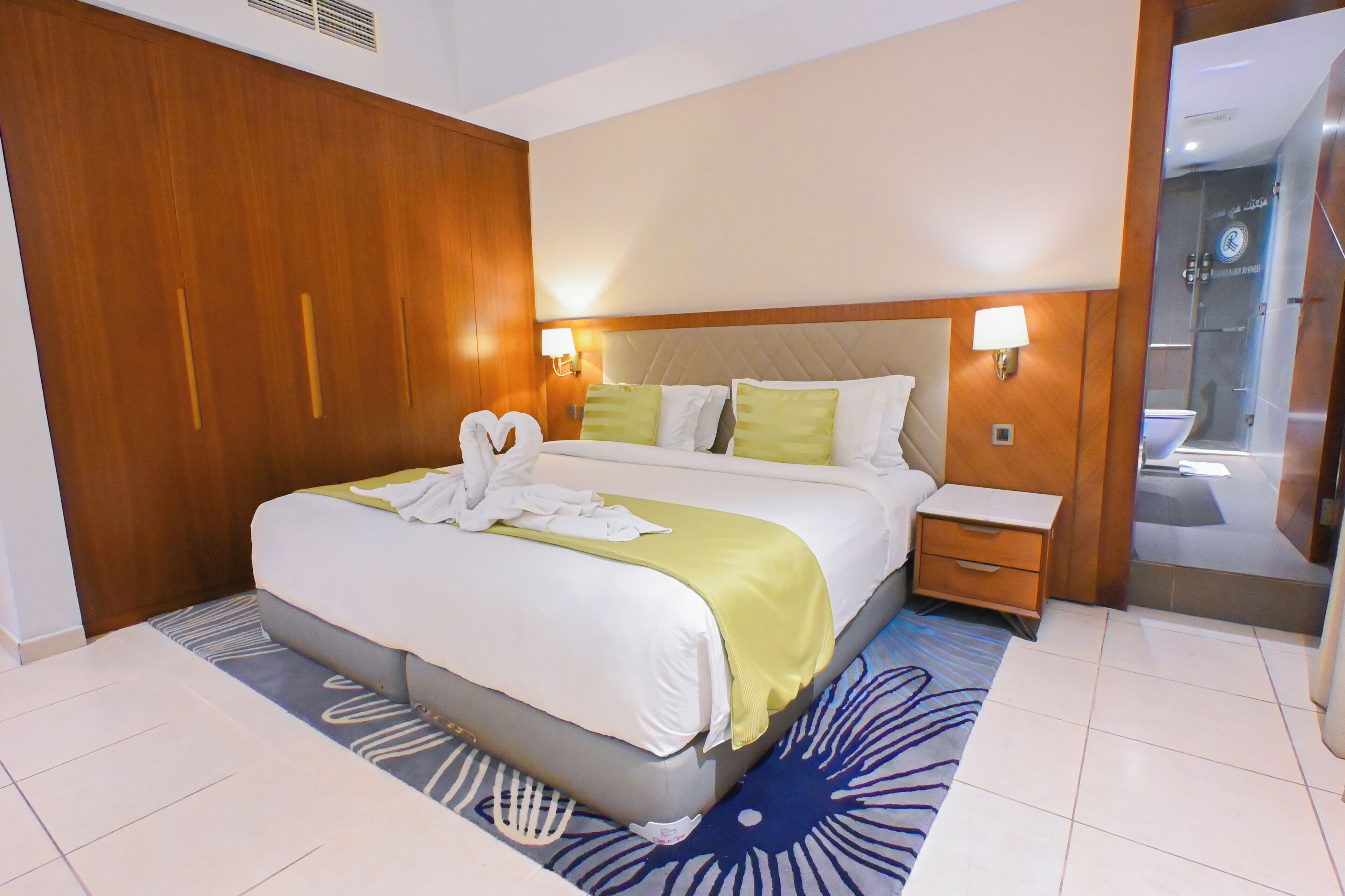 Habitación de hotel con una cama grande, armario de madera, lámparas de noche y baño visible a través de una puerta de cristal.