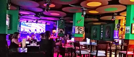 Un restaurante con un interior vibrante, que cuenta con un escenario para presentaciones en vivo y una zona de comedor informal con mesas y sillas.