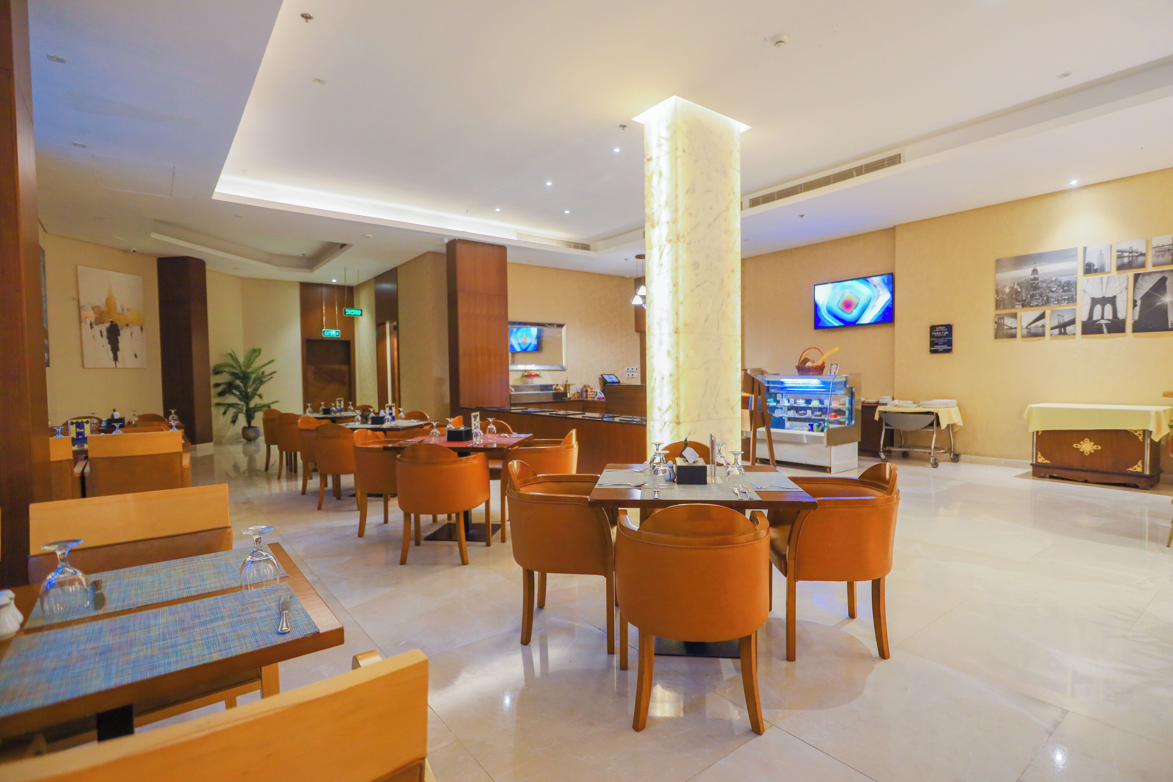 A hotel lobby with multiple dining tables, orange chairs, a reception desk, and a television screen.