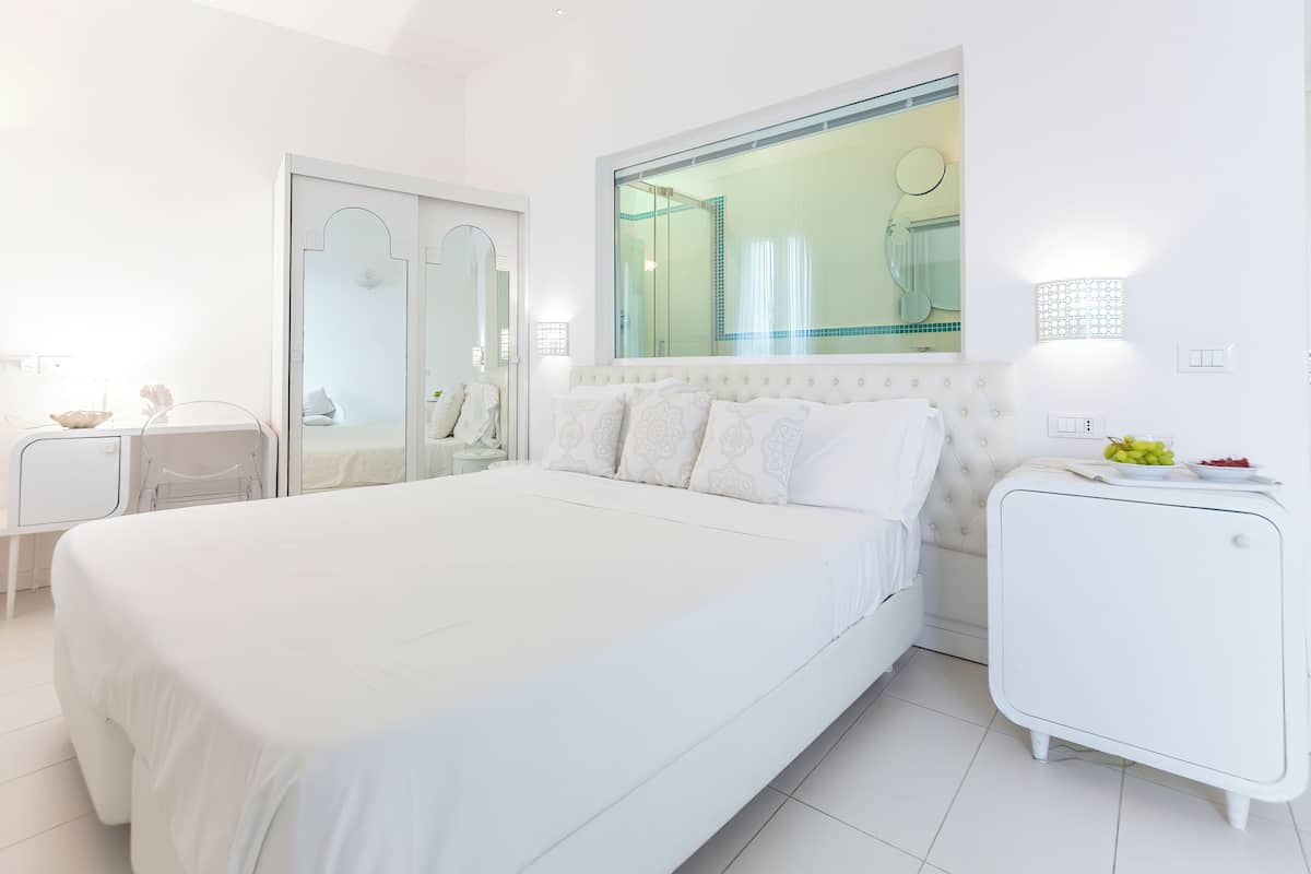 A modern, white-themed bedroom with a large bed, a bedside table, a wardrobe, and a small white cabinet.