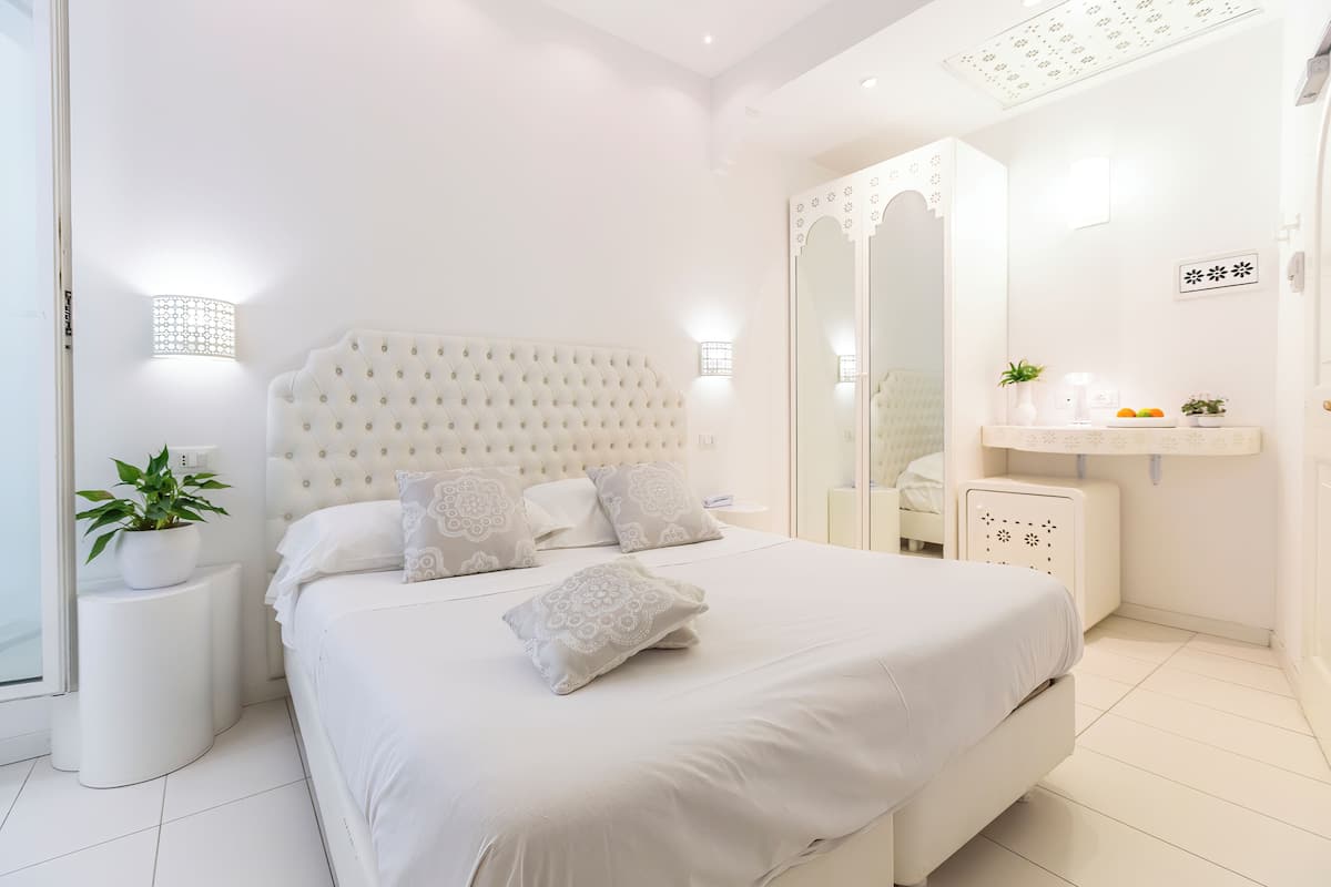 A modern bedroom with a large bed, white bedding, a bedside table with a plant, and a mirror above the dresser.