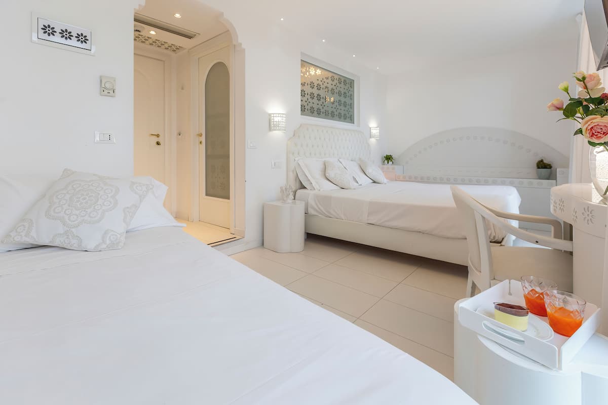 A hotel room with a large bed, a smaller bed, a desk with a chair, and a bedside table with a vase of flowers.