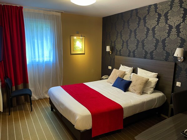A hotel room with a bed, a chair, a bedside table with a lamp, and a wall-mounted telephone.