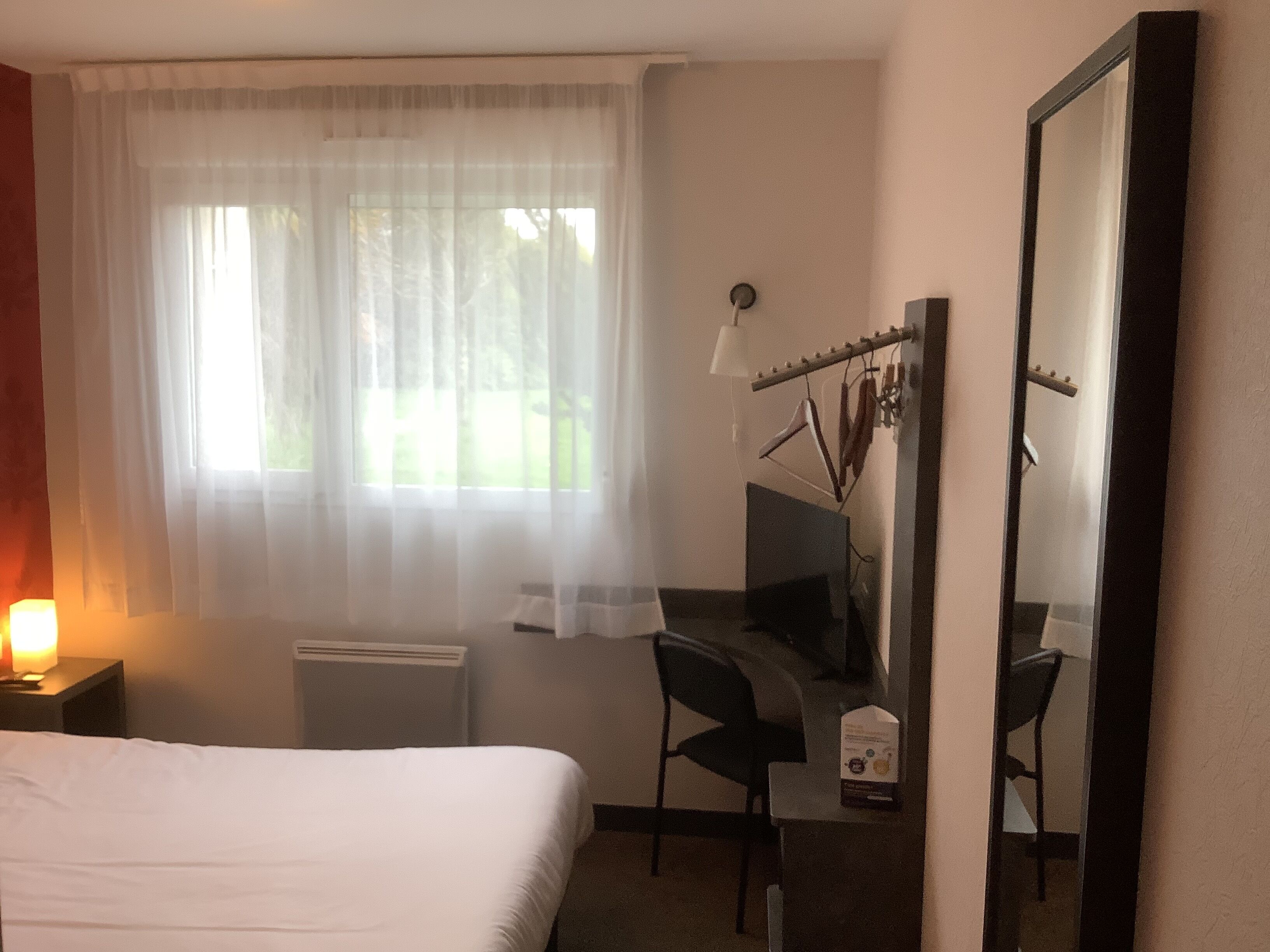 A hotel room with a bed, a desk, a chair, a mirror, and a window with sheer curtains.