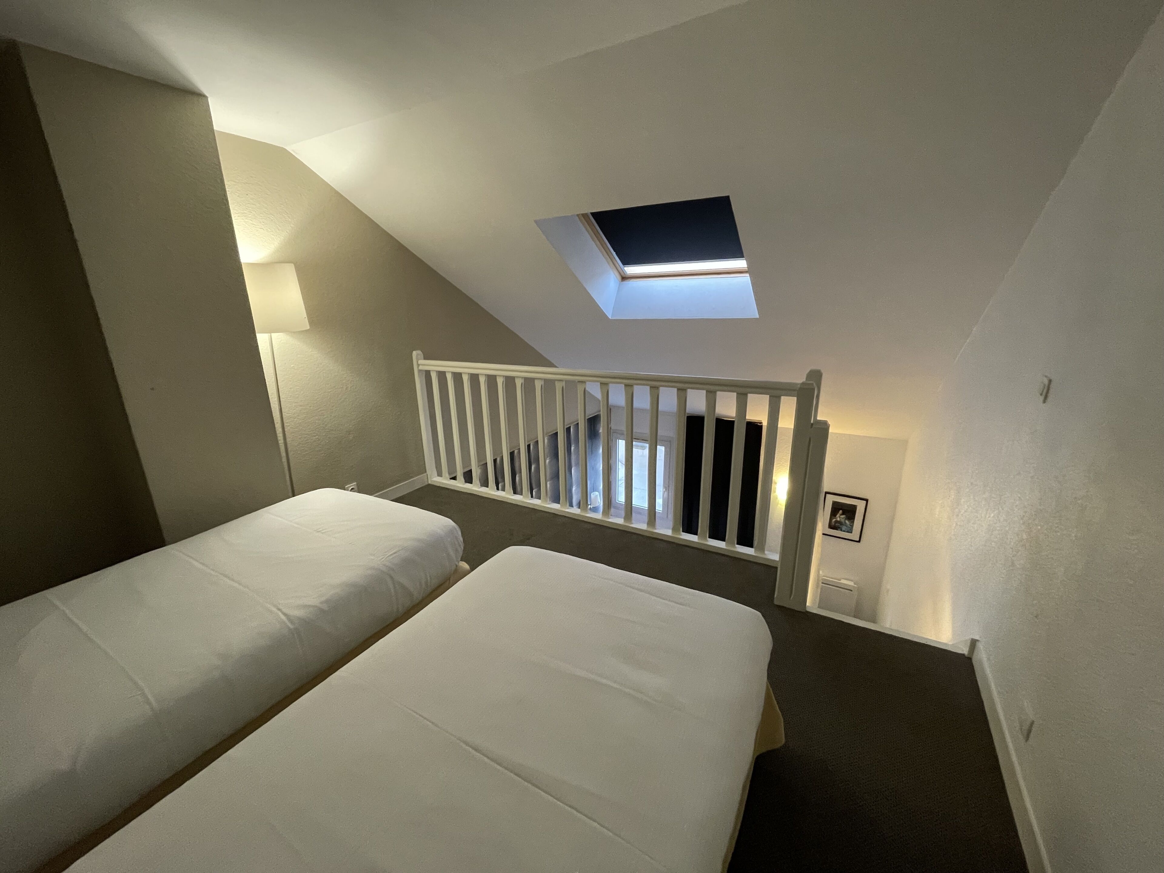 A bedroom with two beds, a skylight, a white railing, and a lamp.