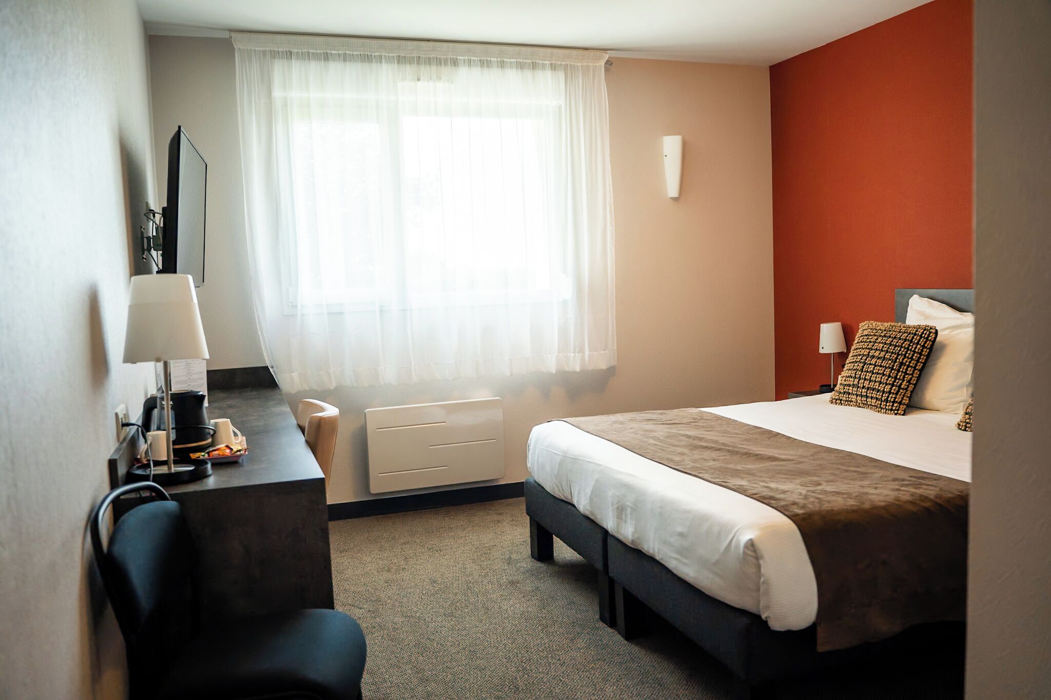 A hotel room with a bed, a desk, a chair, a lamp, a window with sheer curtains, and a wall-mounted TV.