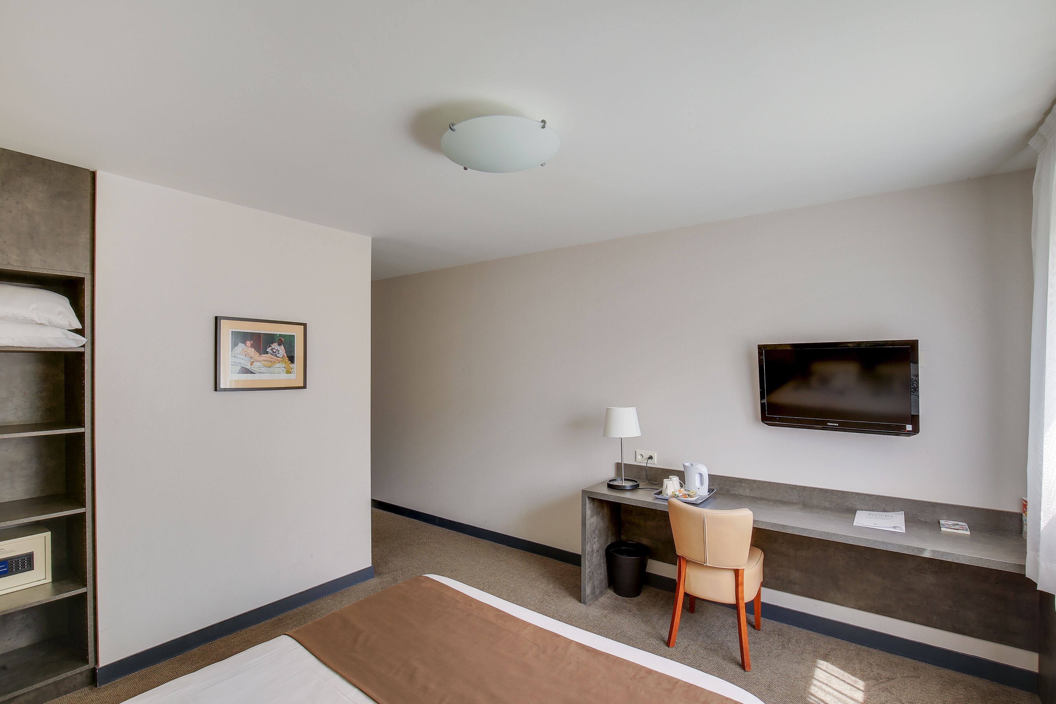 Habitación de hotel con cama, escritorio con silla, televisor, un cuadro en la pared y estante con almohadas.