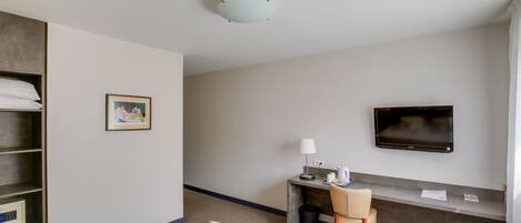 Habitación de hotel con cama, escritorio con silla, televisor, un cuadro en la pared y estante con almohadas.