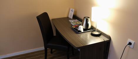 A hotel room with a wooden desk, a chair, a lamp, a kettle, and a tray with a cup and a menu.