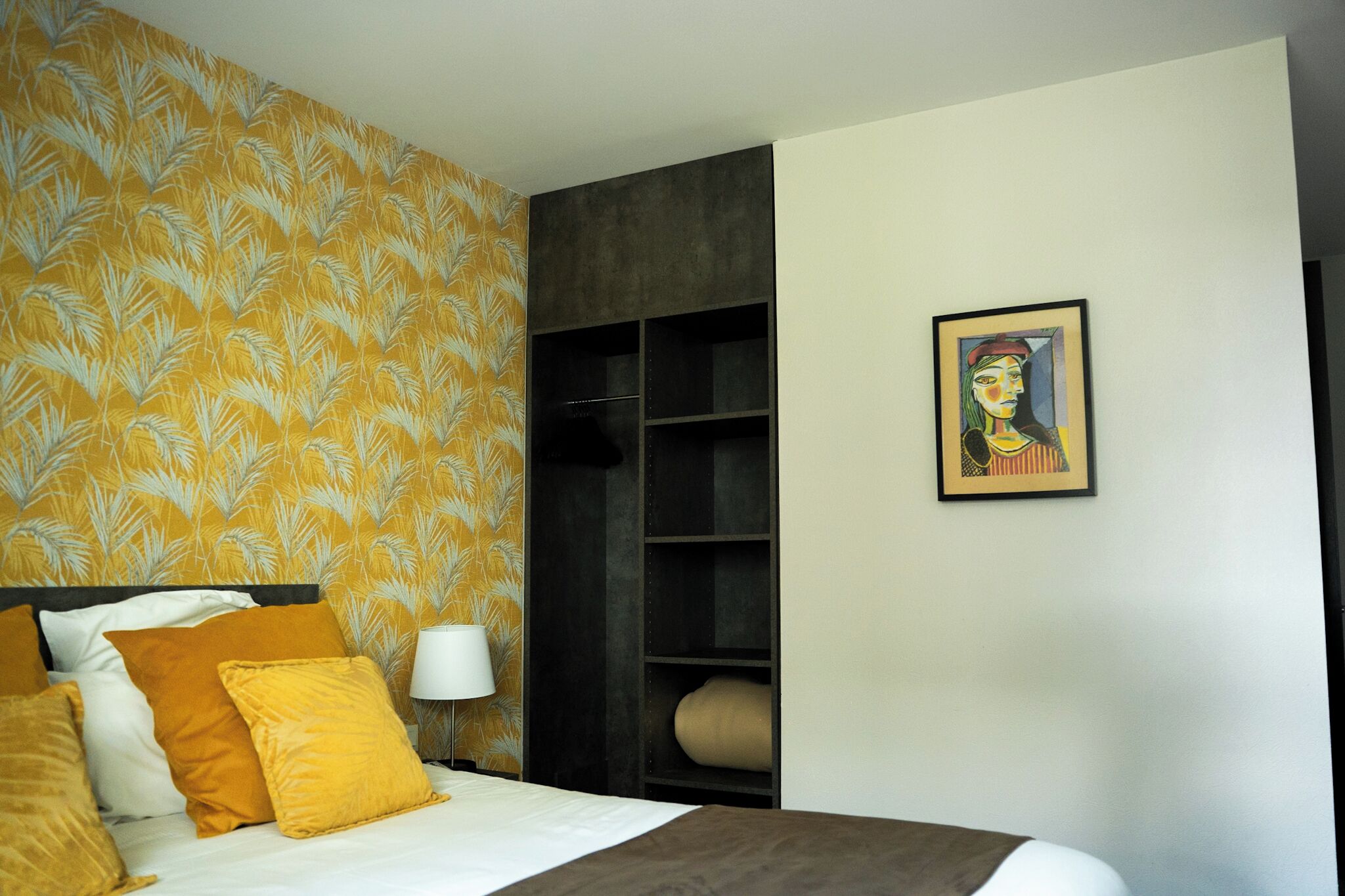 A hotel room with a bed, a headboard, a wall-mounted shelf, and a framed artwork.