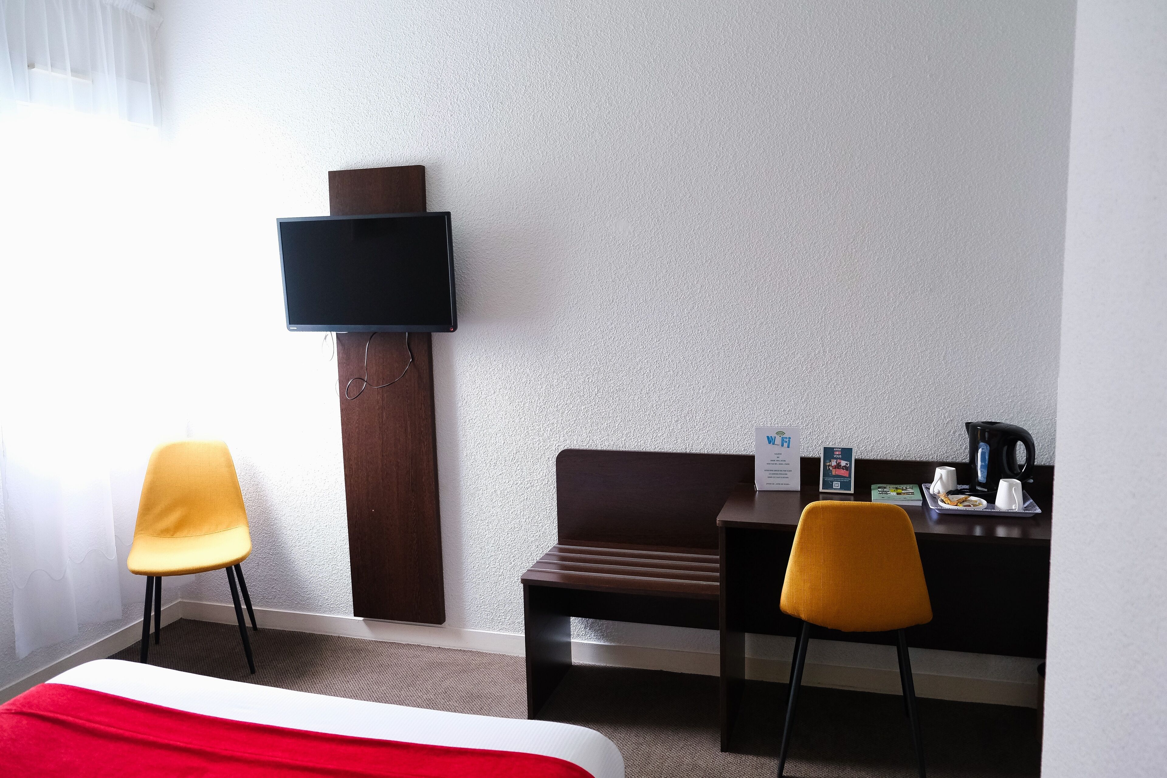 A hotel room with a bed, two chairs, a desk with a coffee maker, and a flat-screen TV mounted on the wall.