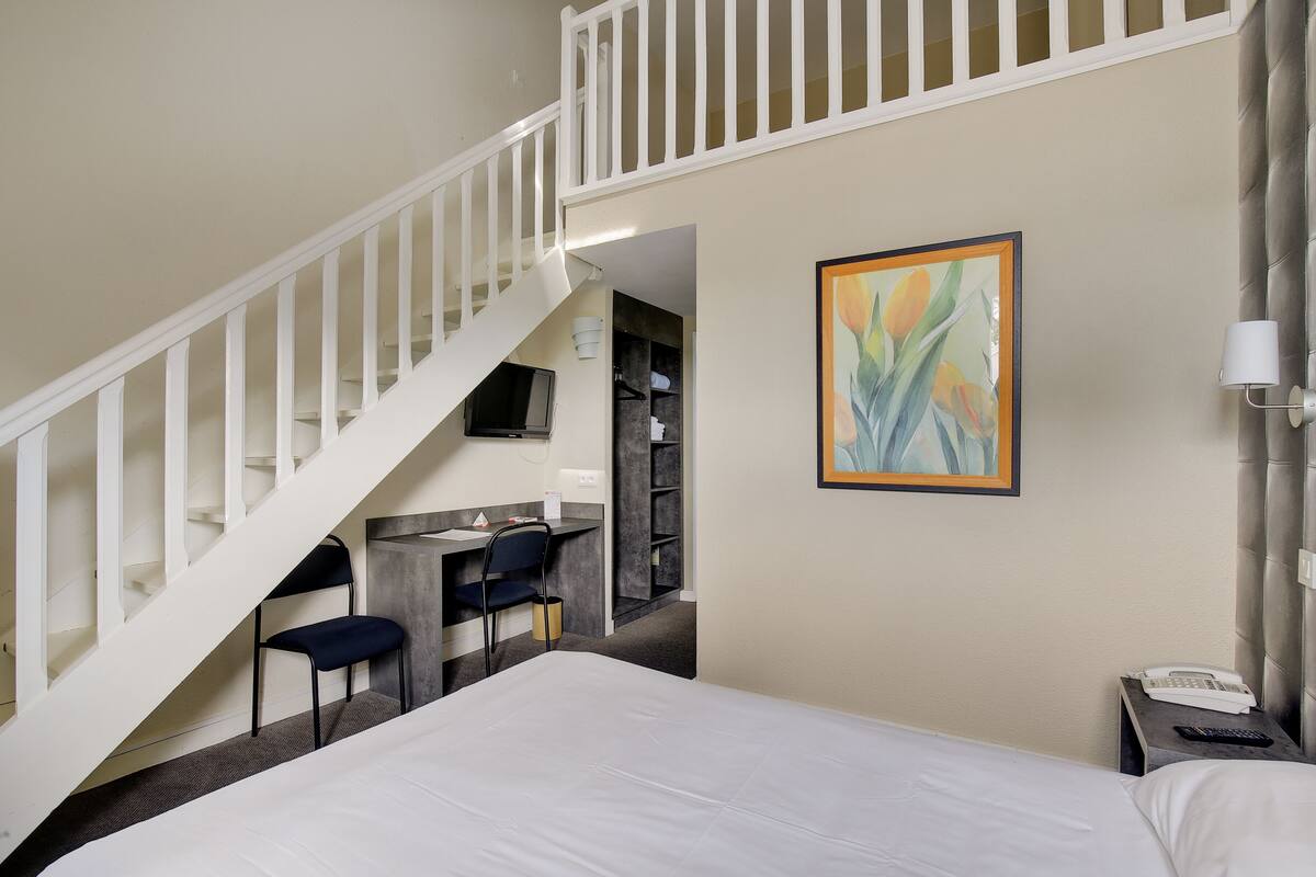A hotel room with a bed, a desk with a chair, a painting, and a staircase.