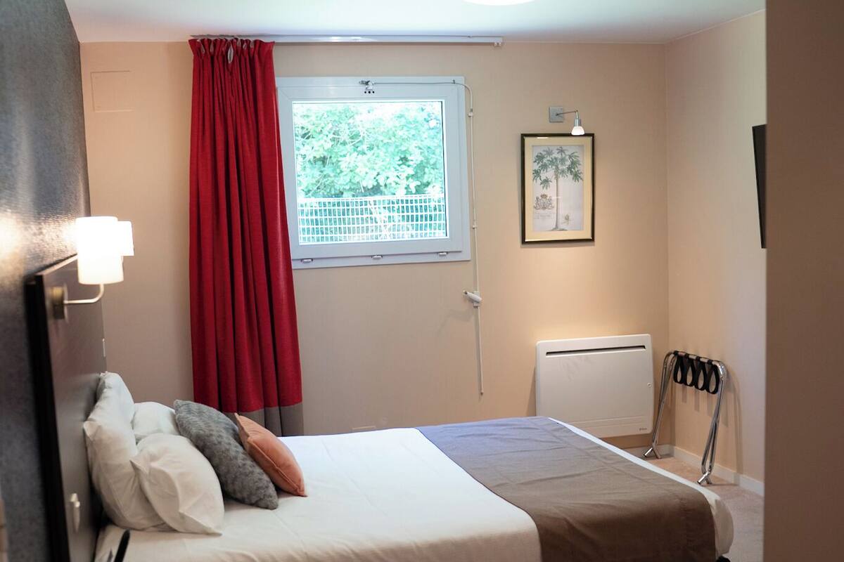 A bedroom with a bed, a window with red curtains, a radiator, and a framed picture on the wall.