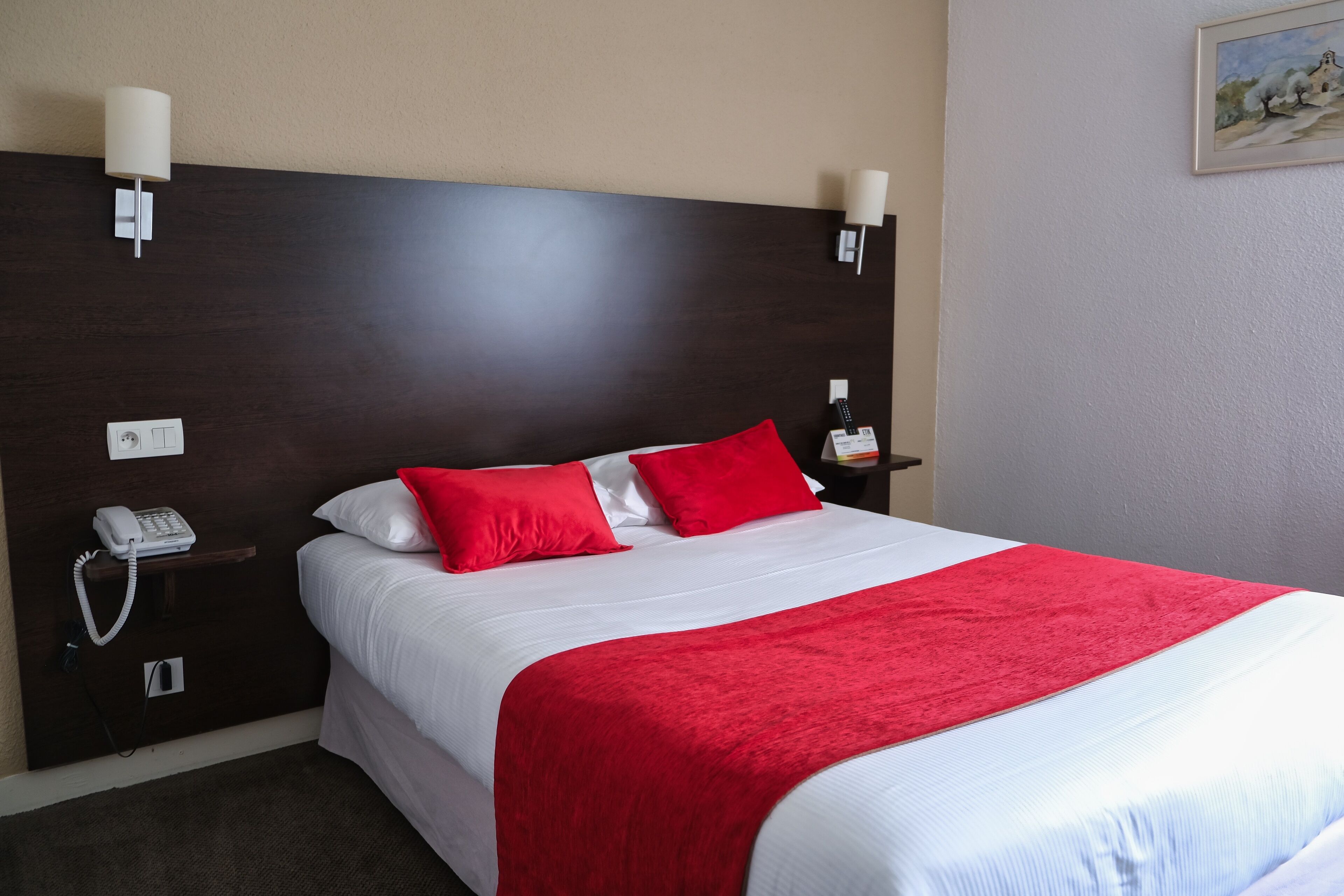 A hotel room with a large bed, white bedding, red pillows, a red throw blanket, a bedside table with a lamp, a phone, and a painting on the wall.
