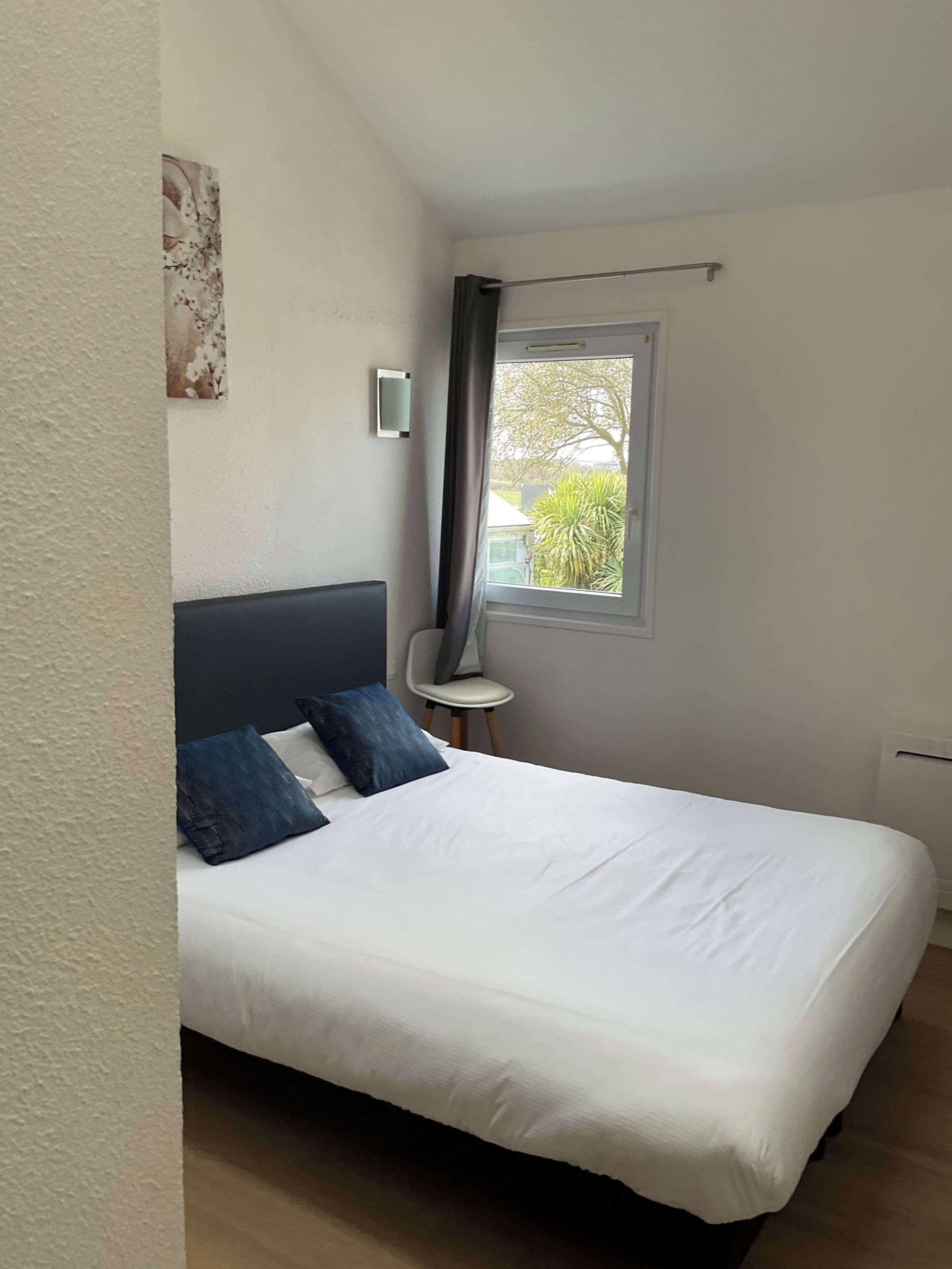 Une chambre à coucher comprenant un lit, une petite table, une chaise, une fenêtre donnant sur un espace verdoyant et un luminaire fixé au mur.