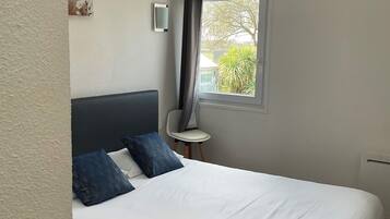 A bedroom with a bed, a small table, a chair, a window with a view of greenery, and a wall-mounted light.