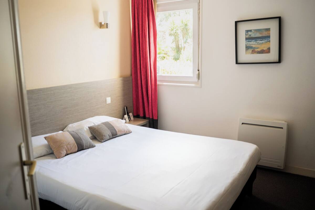 A neatly made bed with pillows, a window with a view, a framed picture on the wall, and a radiator.