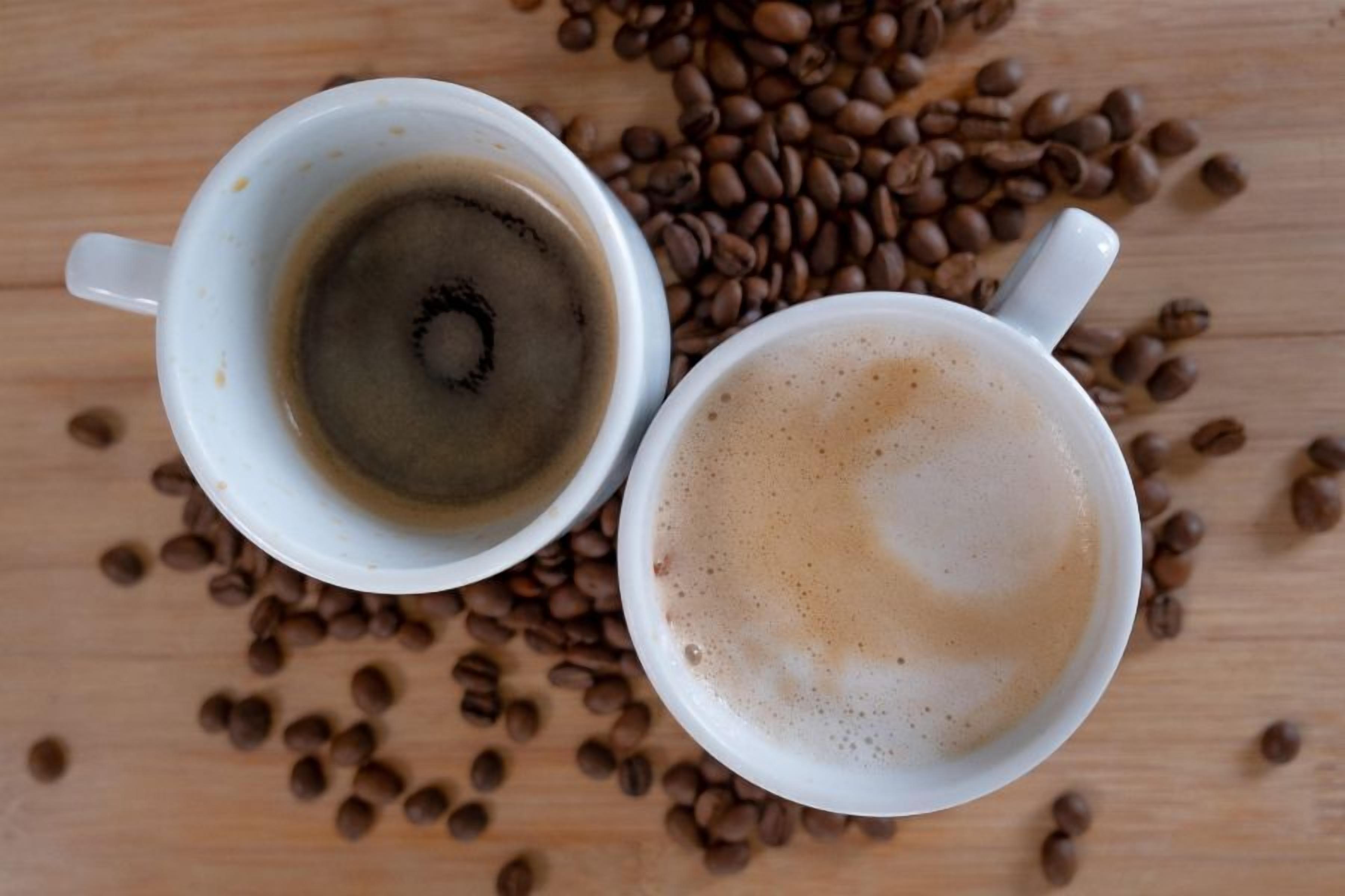 Two white cups of coffee on a wooden surface with scattered coffee beans.