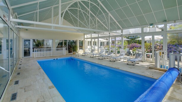 An indoor swimming pool with a glass ceiling, lounge chairs, and a white fence. - Hotel Arcadia (Lannion)