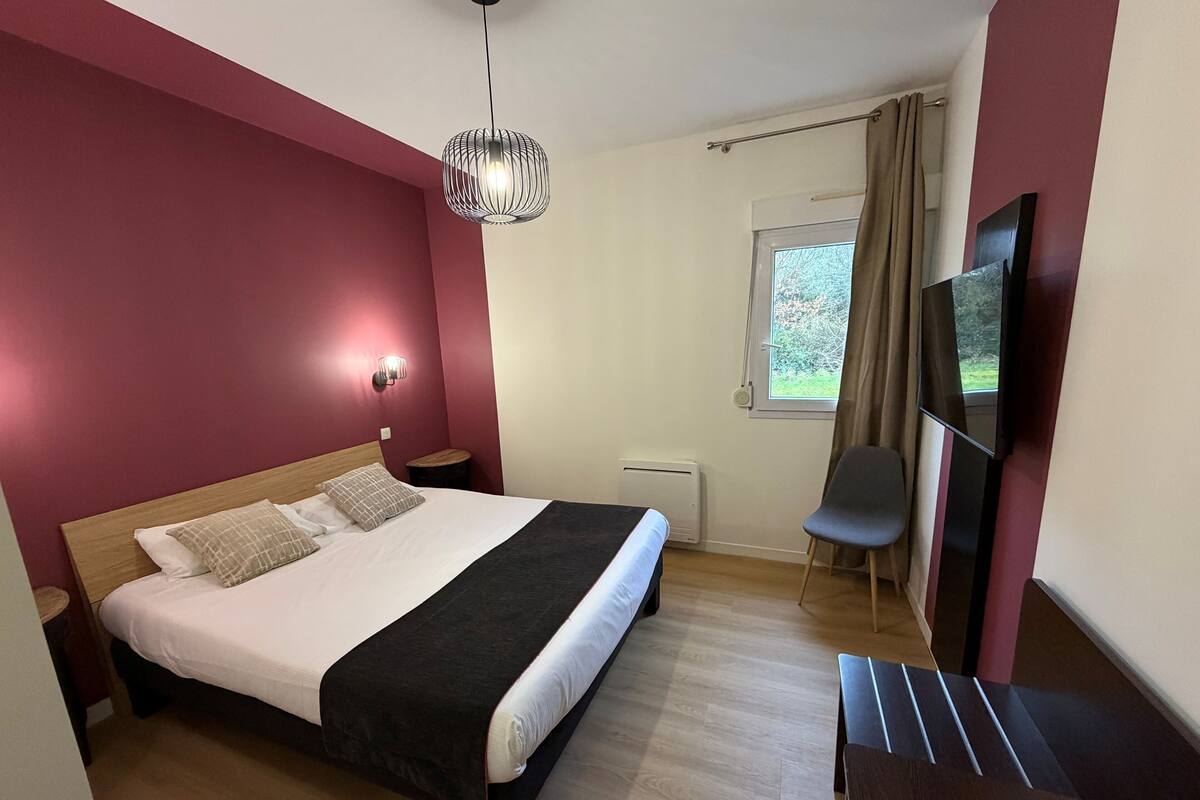 A modern bedroom with a large bed, a bedside table, a chair, a TV, and a window with curtains.
