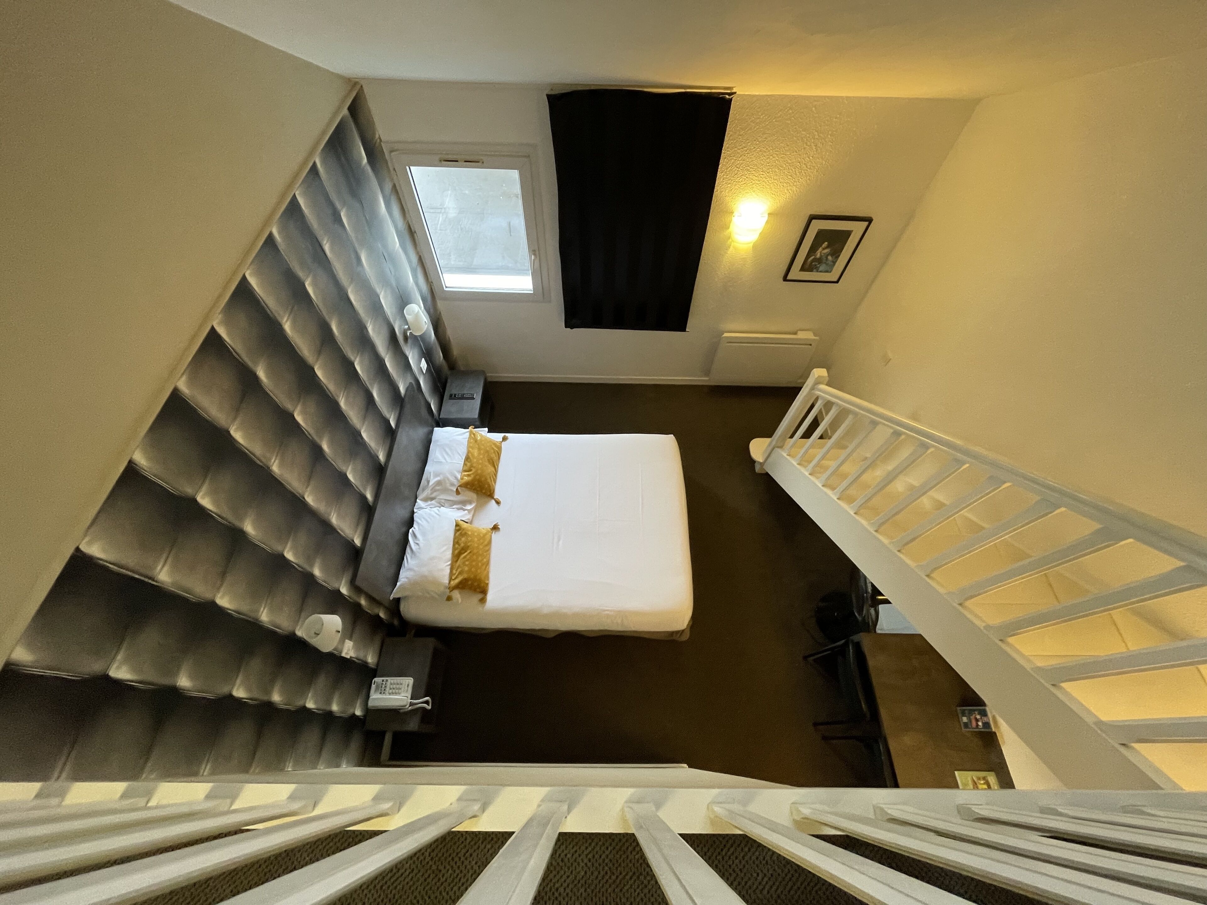 A hotel room with a bed, a nightstand, a window, a picture on the wall, and a staircase.