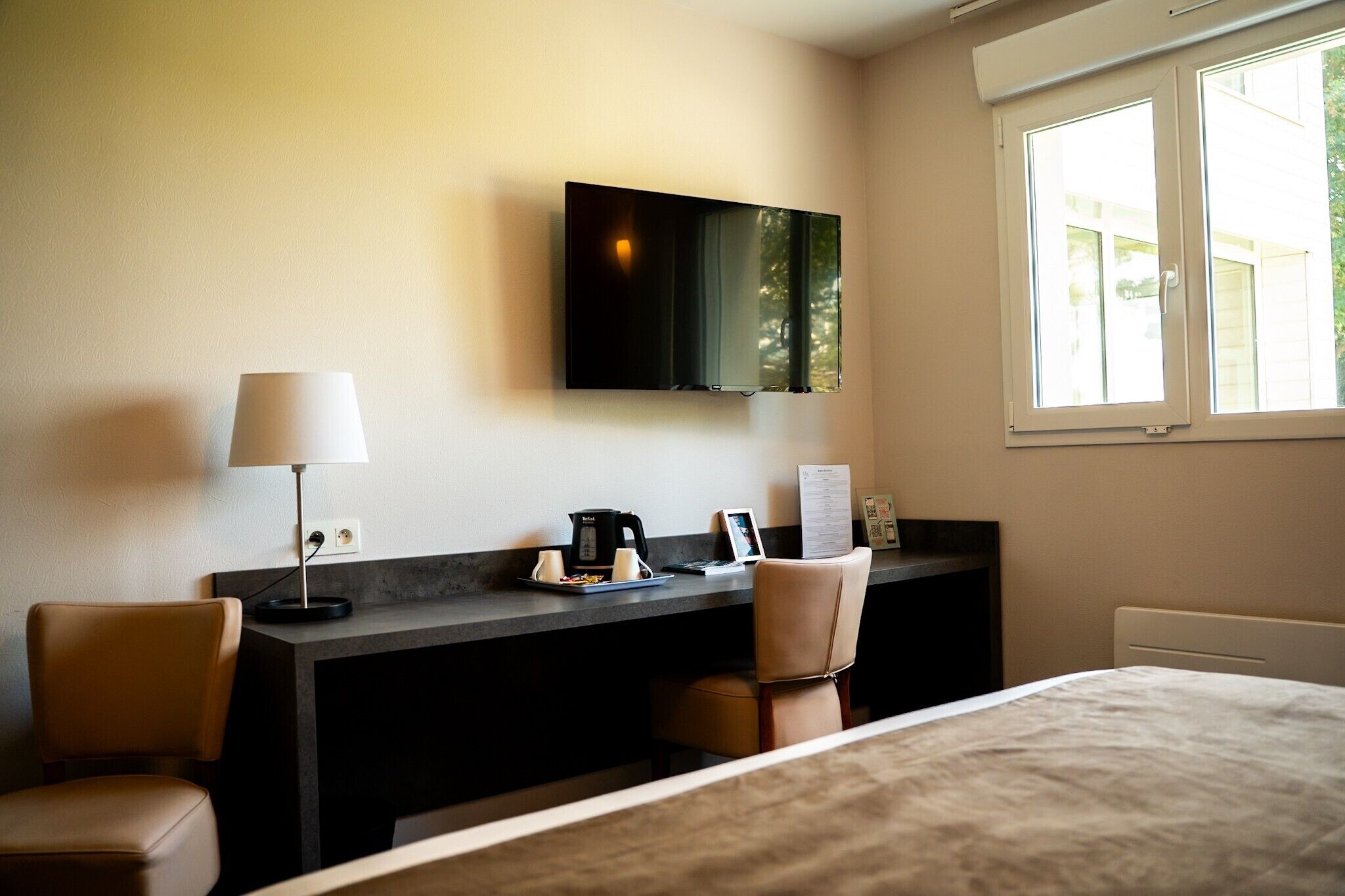 A hotel room with a bed, a desk with a lamp, a flat-screen TV, and a window.