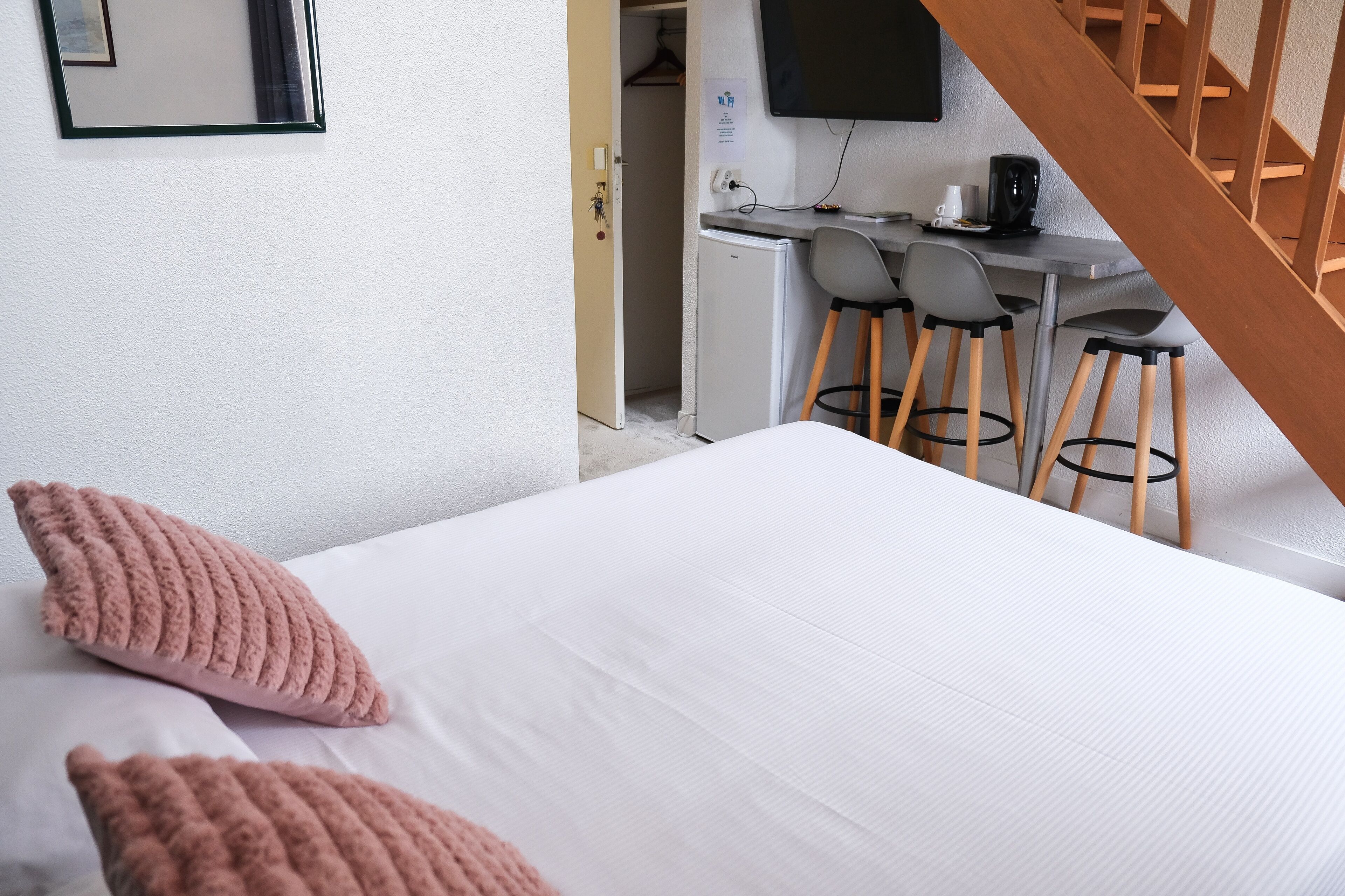 Une chambre d’hôtel avec un lit, deux oreillers roses, un bureau avec un ordinateur, une chaise et un petit réfrigérateur.