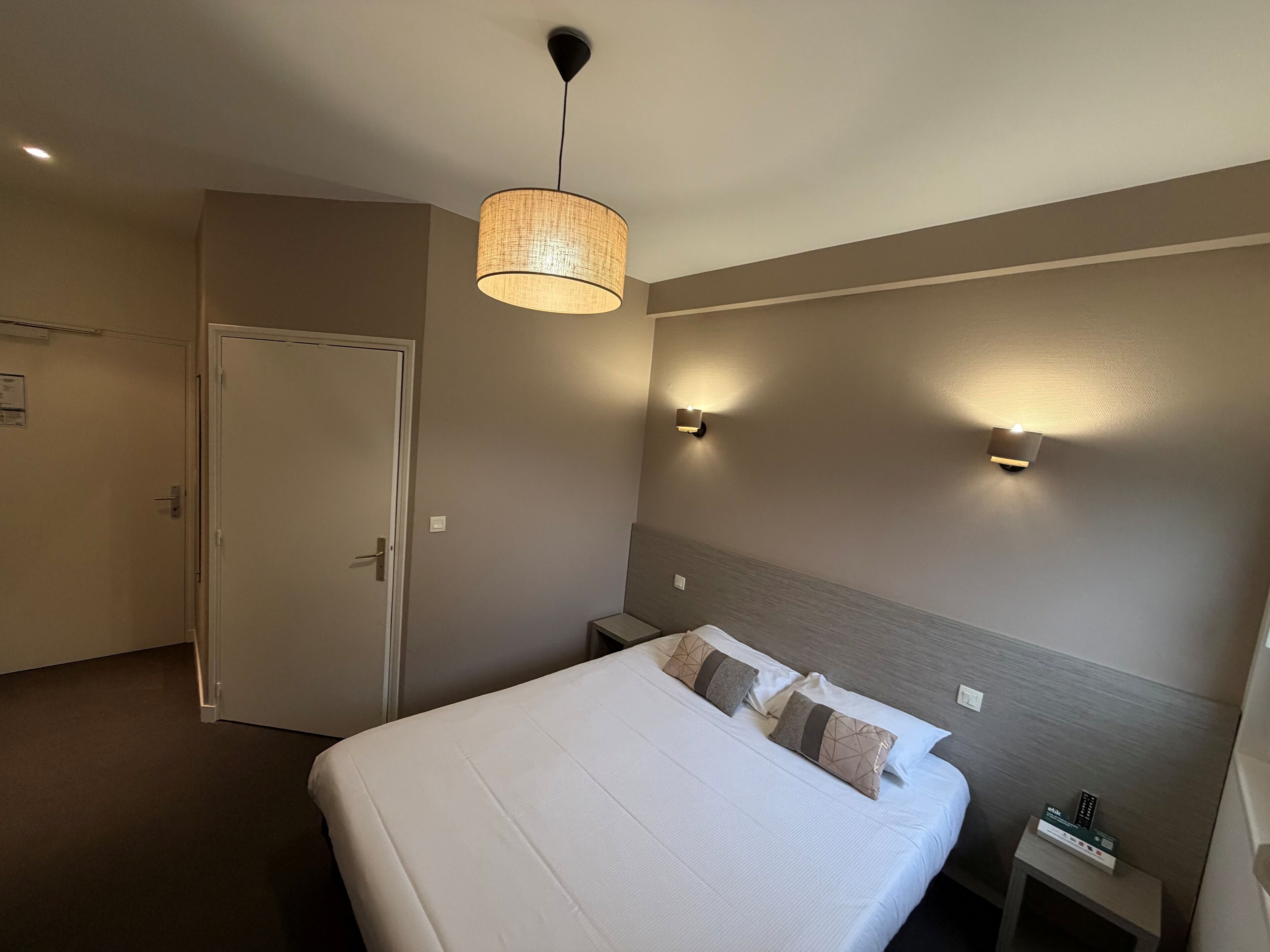 A hotel room with a bed, bedside tables, a hanging lamp, and wall-mounted lights.