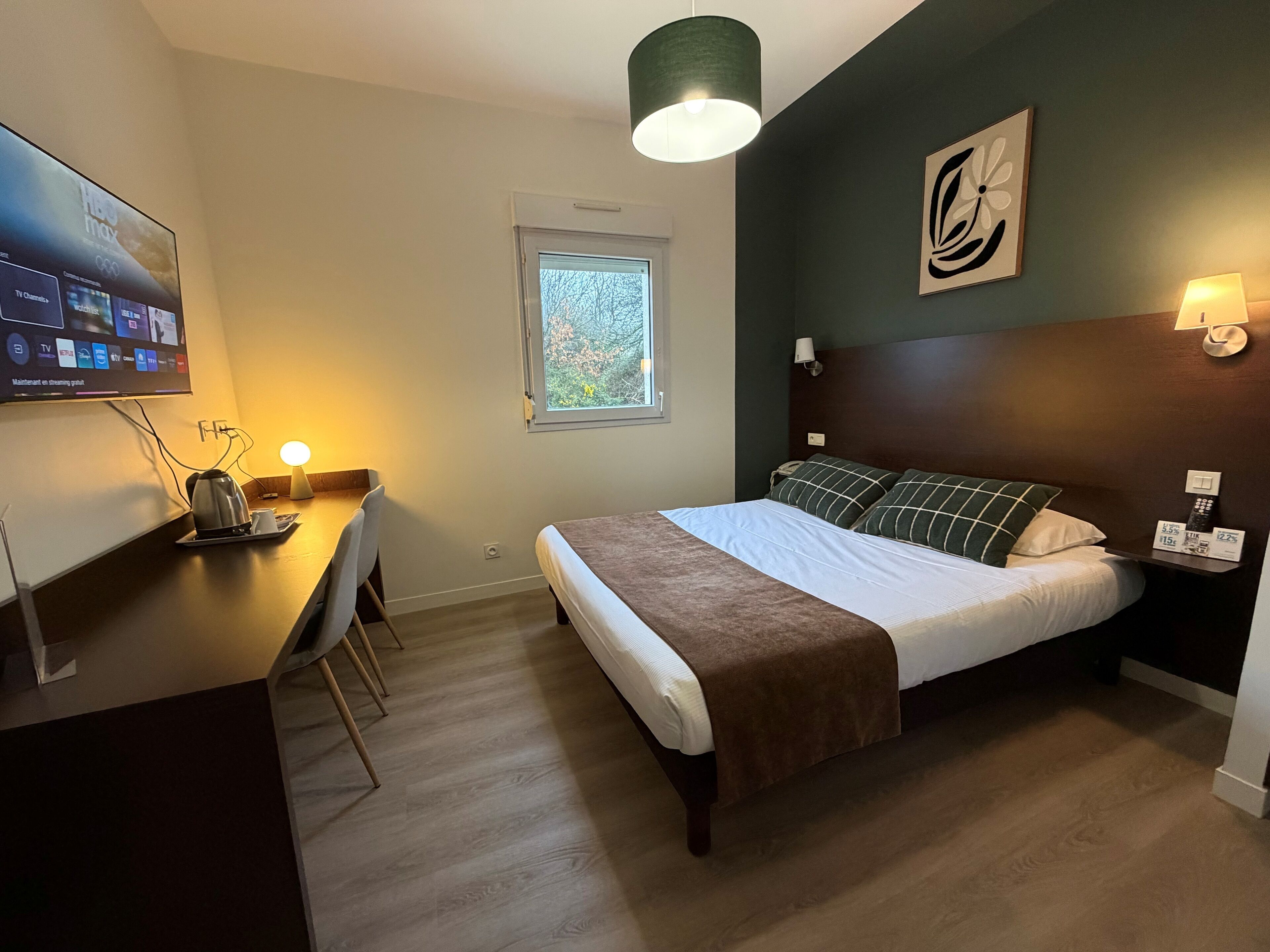 A hotel room with a bed, a desk with a kettle, a TV mounted on the wall, and a window with a view of trees.