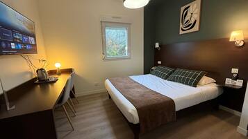 Habitación de hotel con cama, escritorio con hervidor, televisor montado en la pared y ventana con vistas a árboles.
