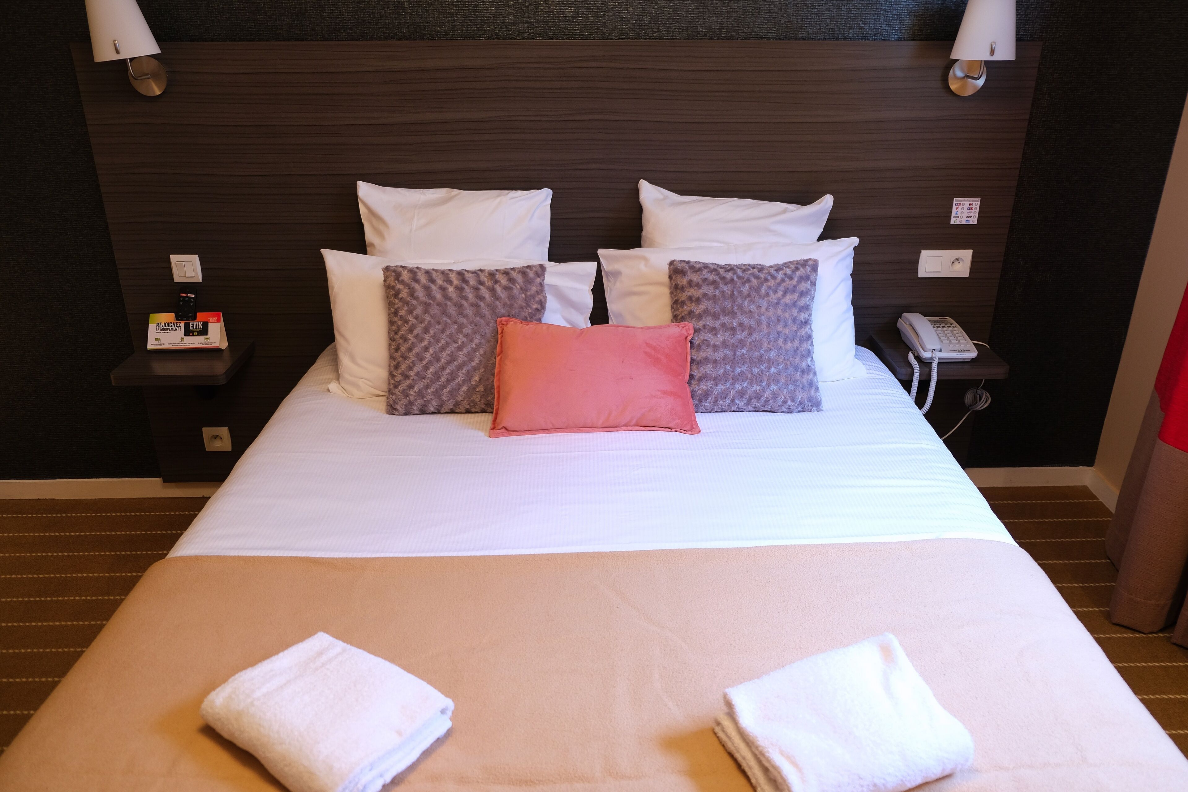 Una cama bien hecha con almohadas blancas y estampadas, una mesita de noche con un teléfono y una lámpara, y una lámpara fijada a la pared.